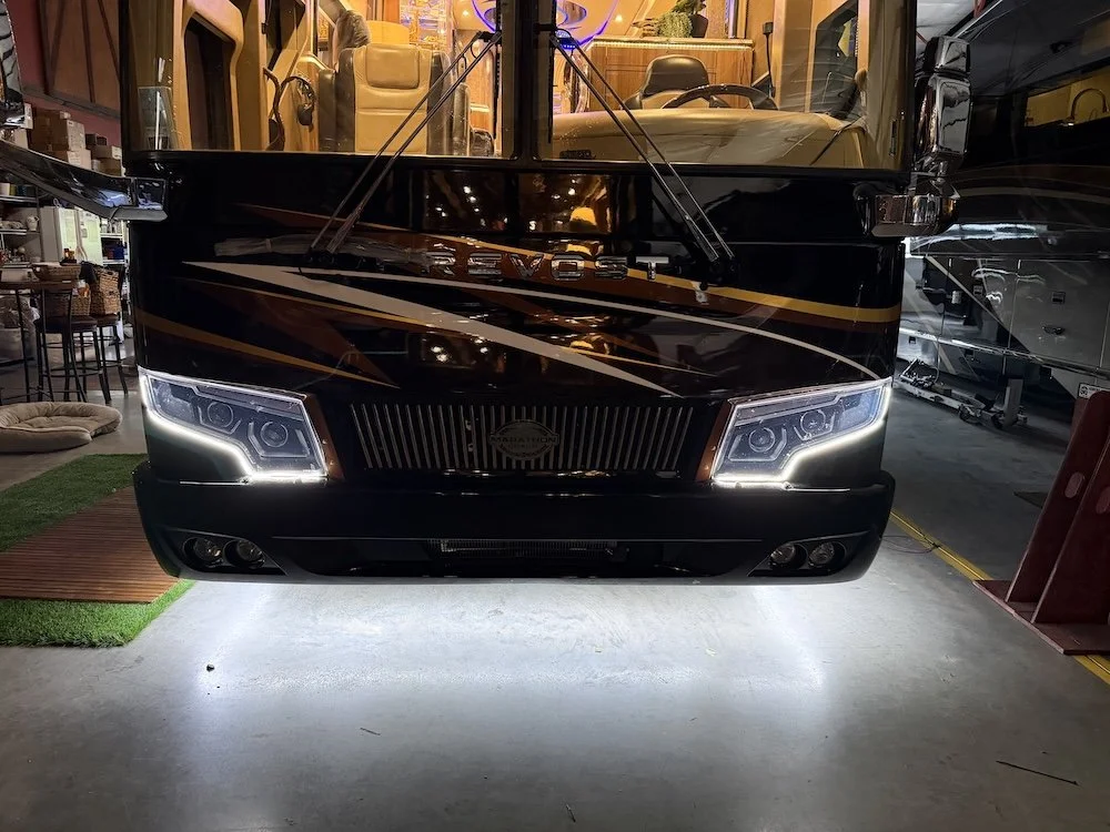 Front view of an RV with illuminated headlights parked inside a showroom or garage.