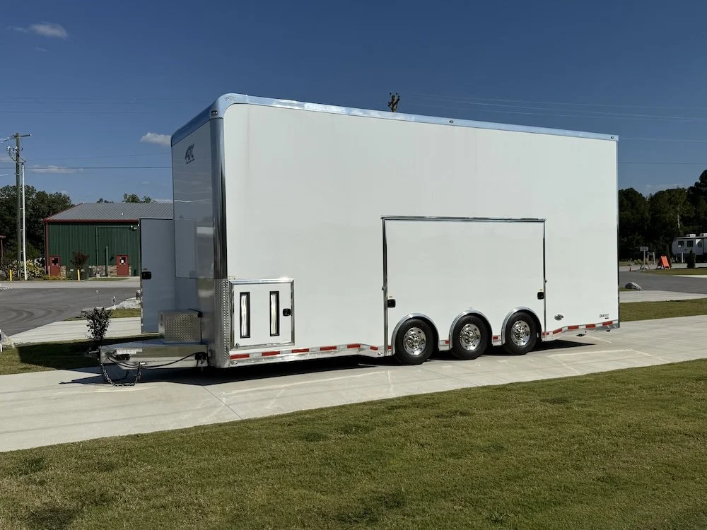 A large white enclosed trailer parked on a concrete driveway with a grassy area in front, under a clear blue sky.