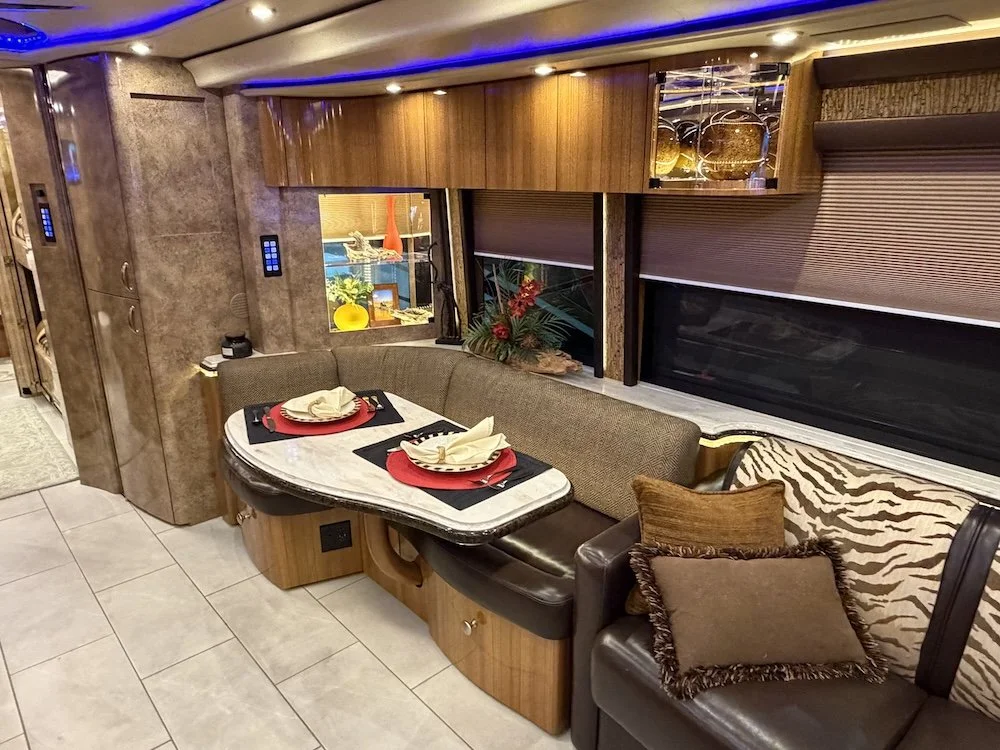 A dining area inside an RV with a marble table set with napkins and plates, a curved sofa with decorative pillows, large windows with closed blinds, and wood cabinetry on the walls.