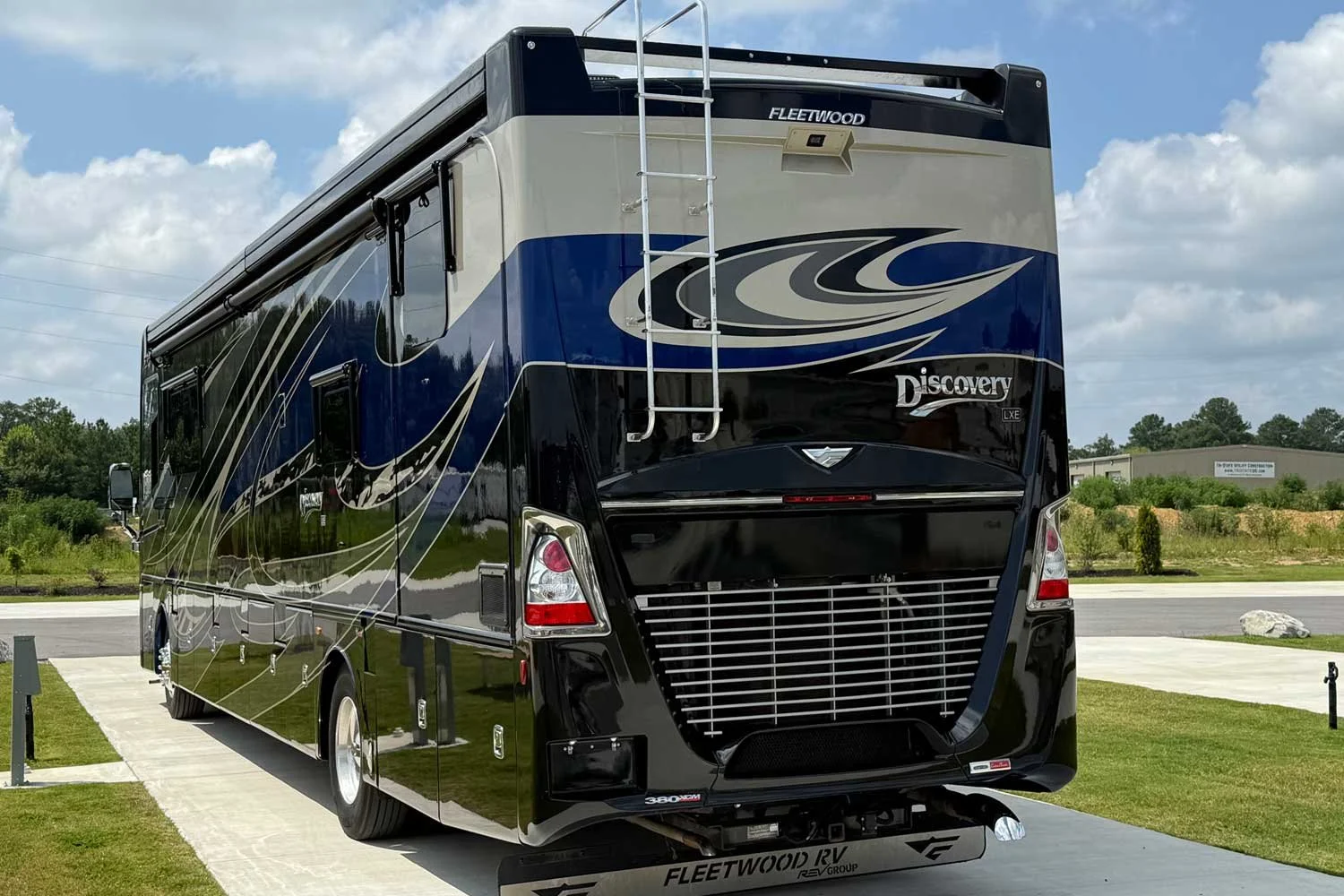 A black and blue Fleetwood Discovery RV parked on a concrete pad, with a ladder attached to the back and an air conditioning unit on top, under a partly cloudy sky.