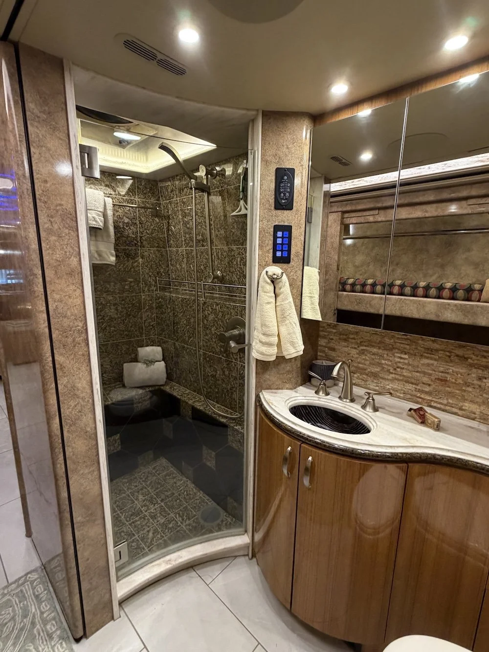 Small bathroom with a corner shower, wooden vanity with a marble top, and a mirror. The shower has brown tiles and a glass door, with towels hanging on a hook outside. 