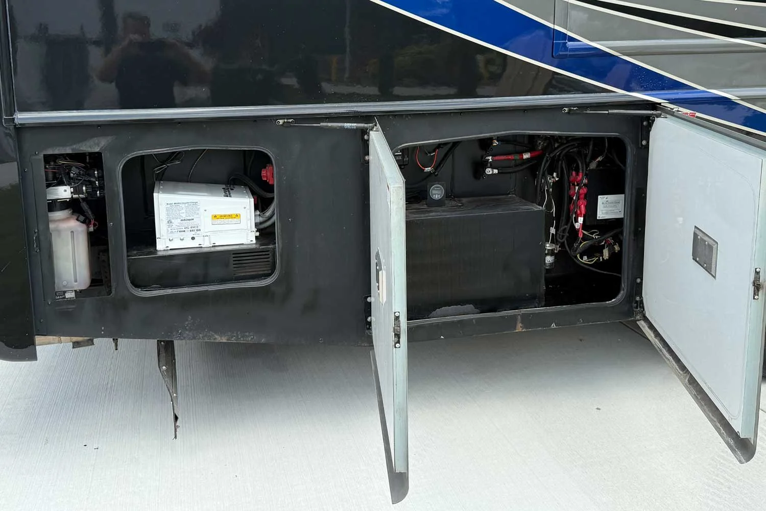 Open compartments showing a battery, electrical components, and wiring inside a recreational vehicle or bus.