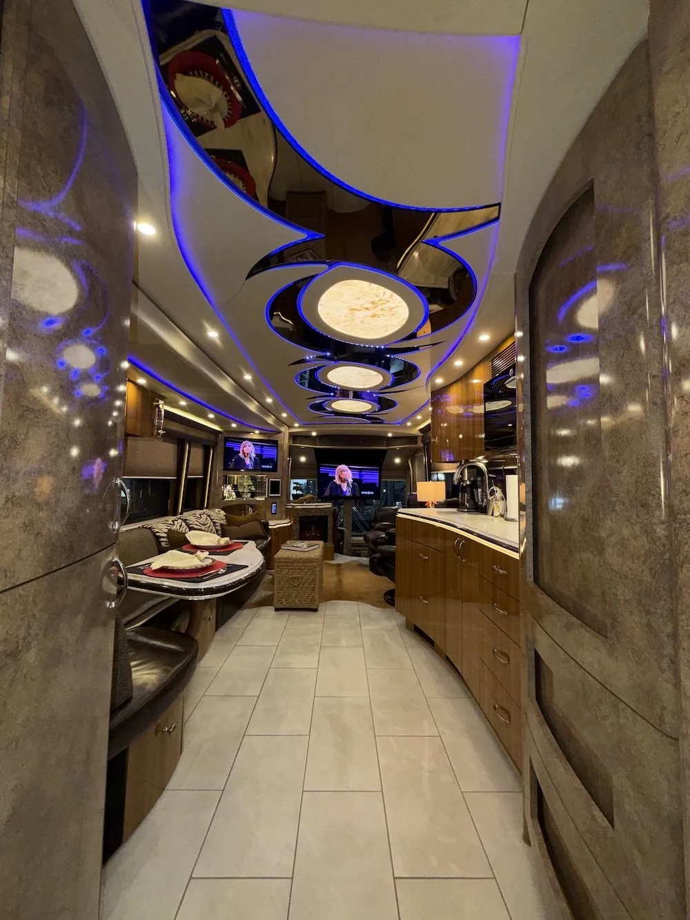 Interior of an RV with modern lighting, a dining or sitting area, and a kitchen counter, illuminated with blue LED lights on the ceiling.