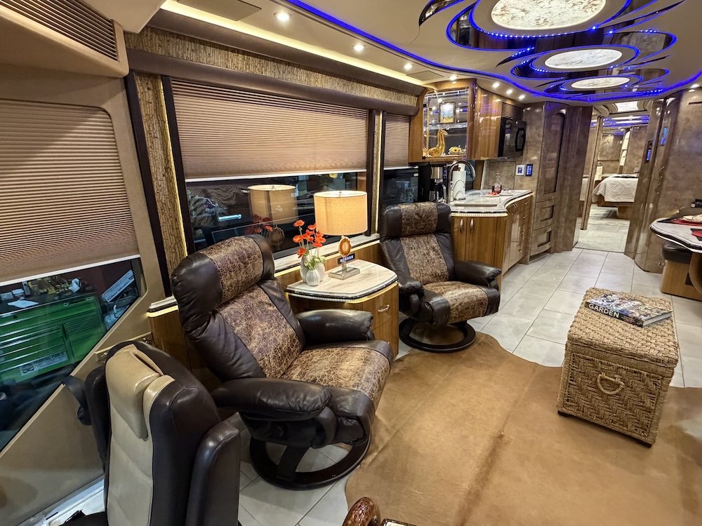 Interior of a luxury RV or motorhome with leather and fabric chairs, a small side table with a lamp and flowers, a woven ottoman with magazines, a kitchenette, and a bedroom in the background.