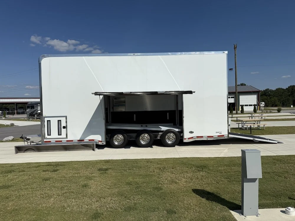 Empty white trailer with side ramp open, parked on a sidewalk in a park-like area under a clear blue sky.