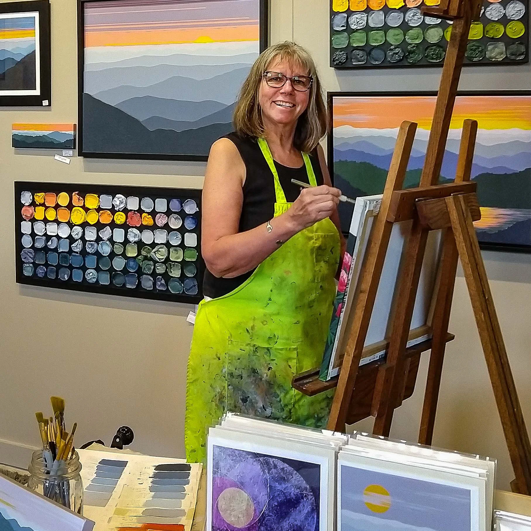 Cindy Chenard in a black top and green apron painting on a canvas at her art gallery/studio. There are landscape paintings on the wall behind her and art supplies on a table in front of her.