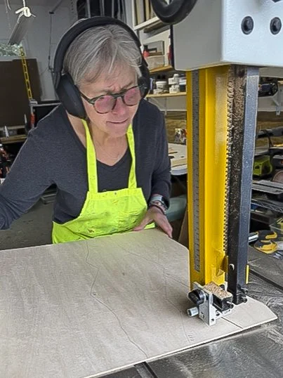 Cindy Chenard wearing a green apron and glasses working with a vertical bandsaw cutting wood for a dimensional landscape painting in her studio workshop.