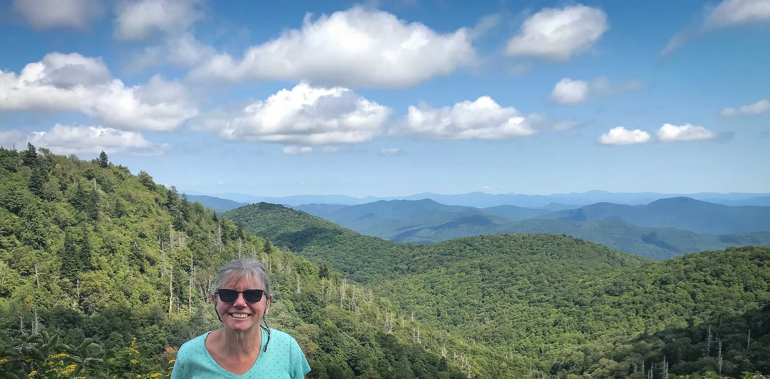 Cindy Chenard wearing sunglasses and a light blue shirt standing on a mountain with green forest and distant blue mountain ridges under a partly cloudy sky.
