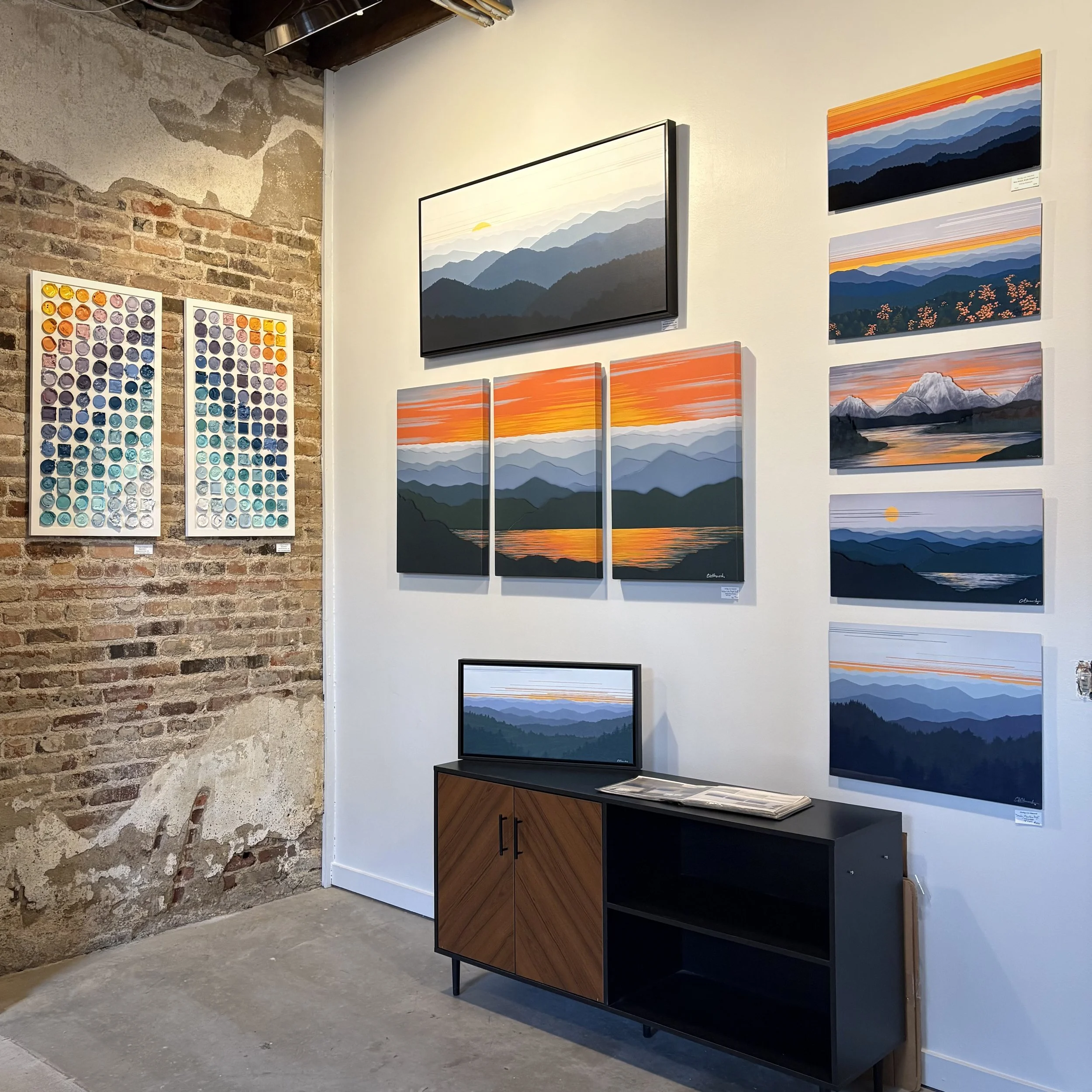 Gallery wall with landscape paintings of mountains and sunsets, a color palette display on a brick wall, and a wooden cabinet with a landscape digital display on top.