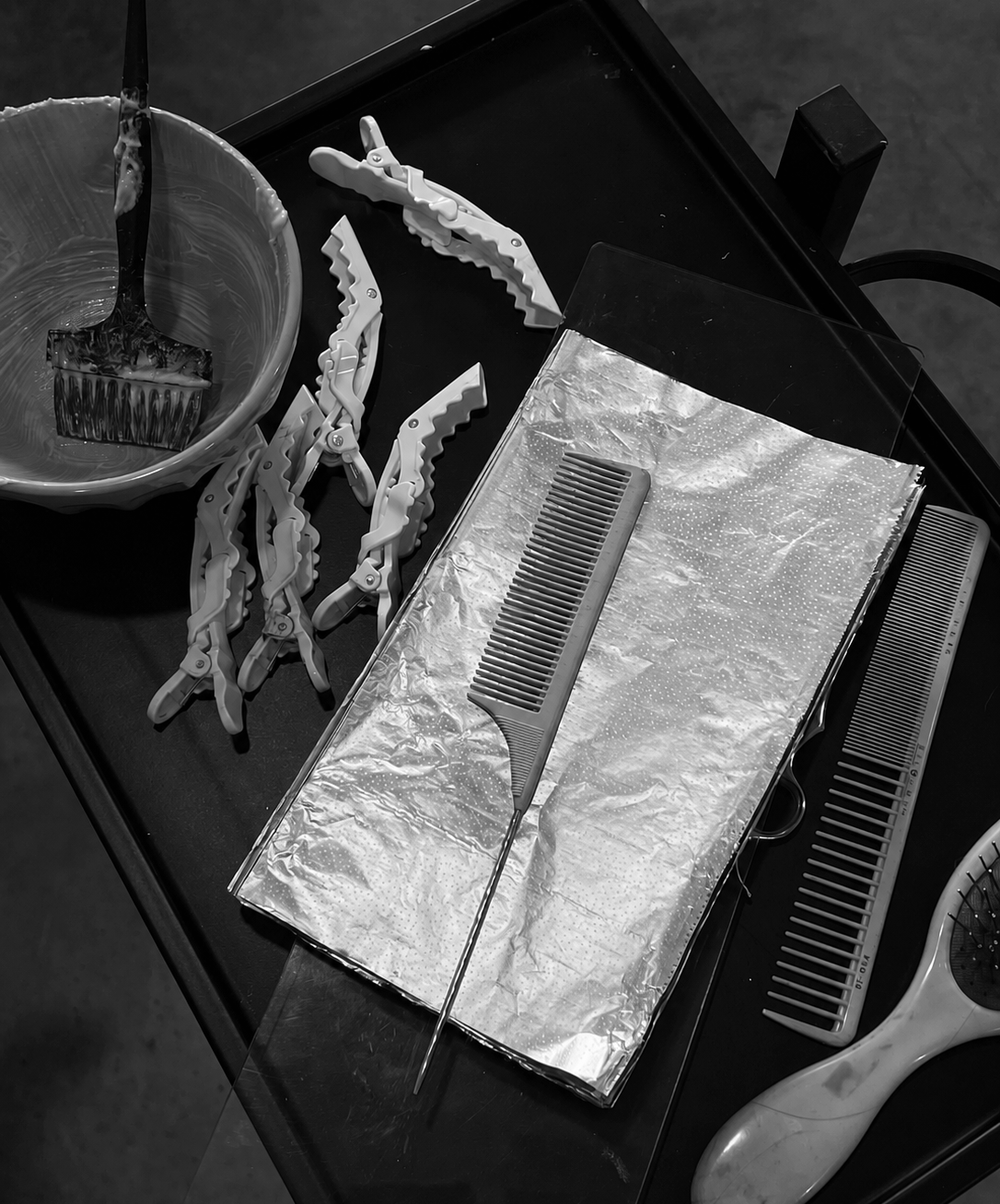 Hair styling tools including clips, a wide-tooth comb, a fine-tooth comb, a brush, and foil sheets are arranged on a black tray.