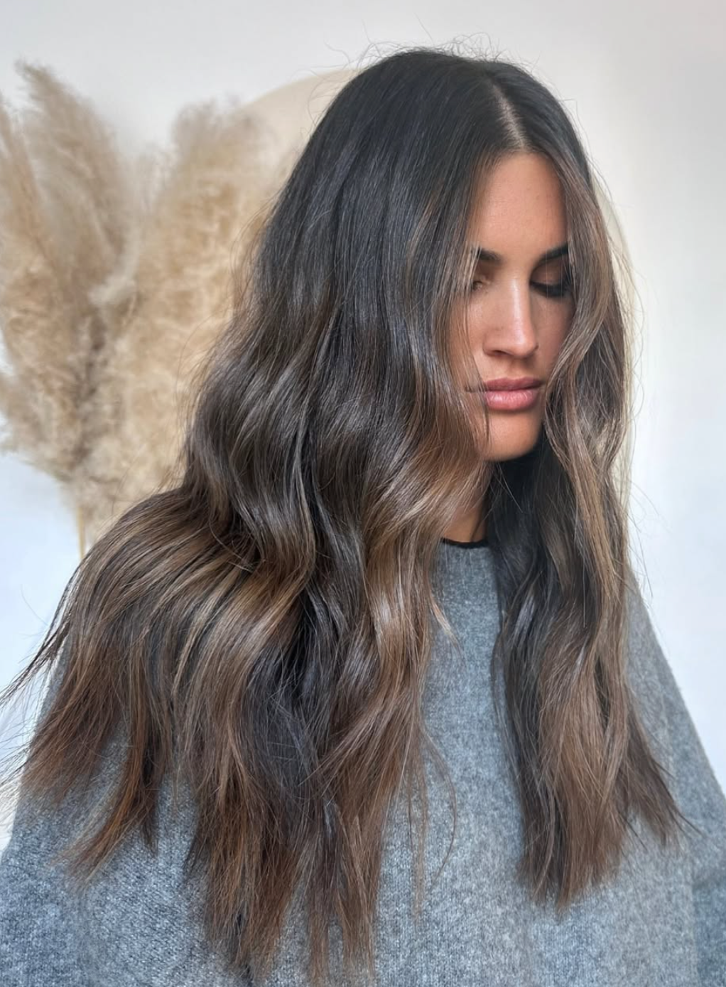 A woman with long, wavy brown hair and light highlights, with her eyes closed and wearing a gray top.