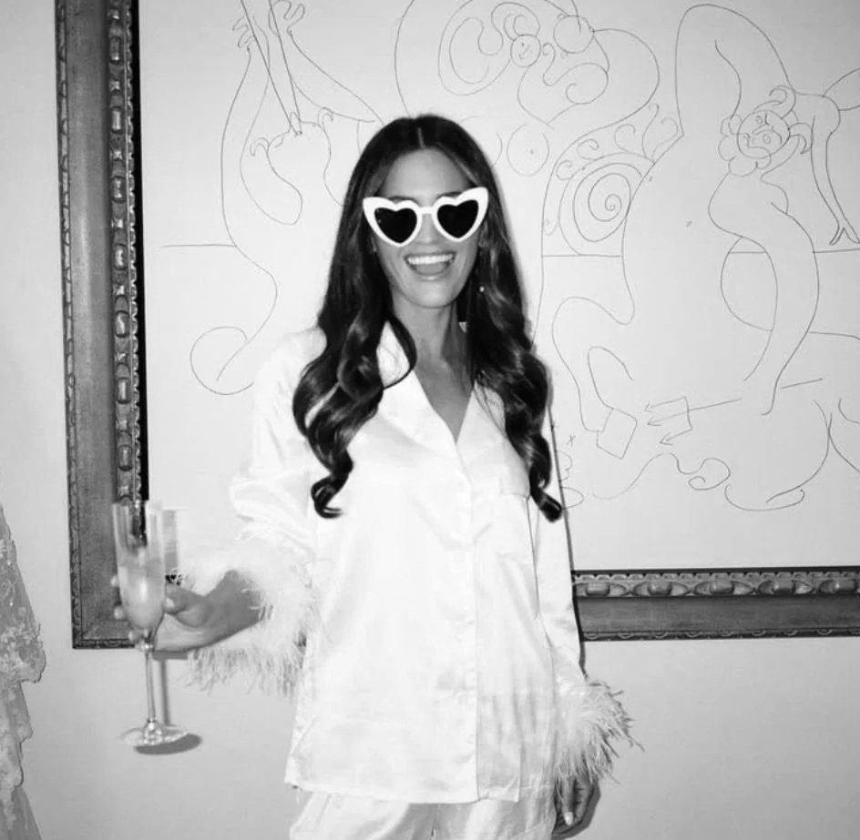 A woman with long wavy hair wearing white pajamas and heart-shaped sunglasses, smiling and holding a glass of champagne, standing in front of a wall with line drawings.