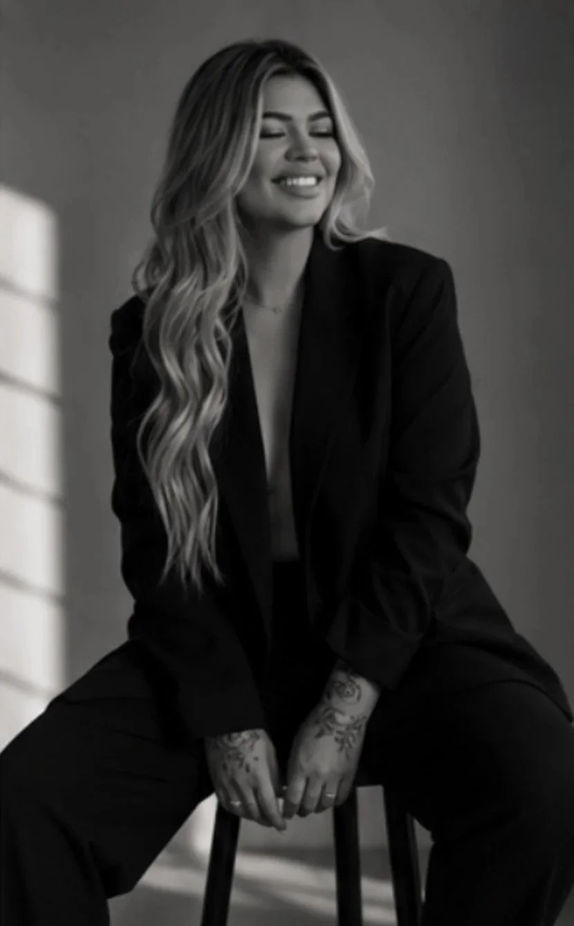 A black and white photo of a woman with long wavy hair, wearing a blazer, smiling with eyes closed, sitting on a stool with her hands resting between her legs, showing tattoos on her hands.