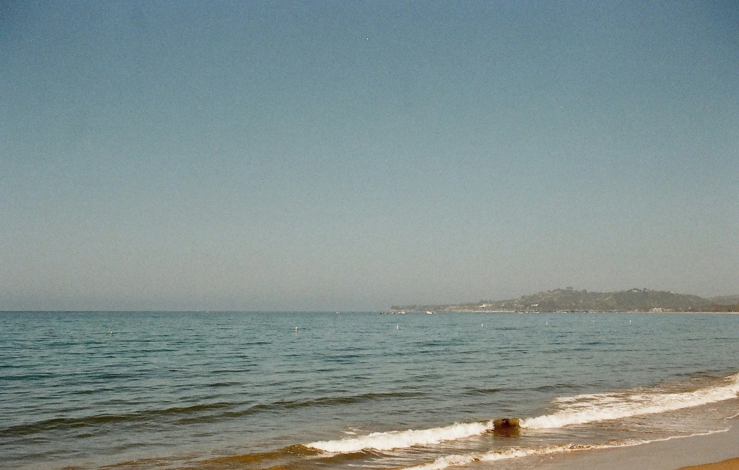 A view of the ocean with gentle waves reaching the sandy shore, under a clear sky with some distant land and low hills in the background.