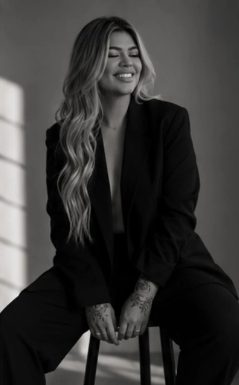 Black and white photograph of a woman with long, wavy hair sitting on a stool, smiling with eyes closed, wearing a dark blazer and displaying tattoos on her hands.
