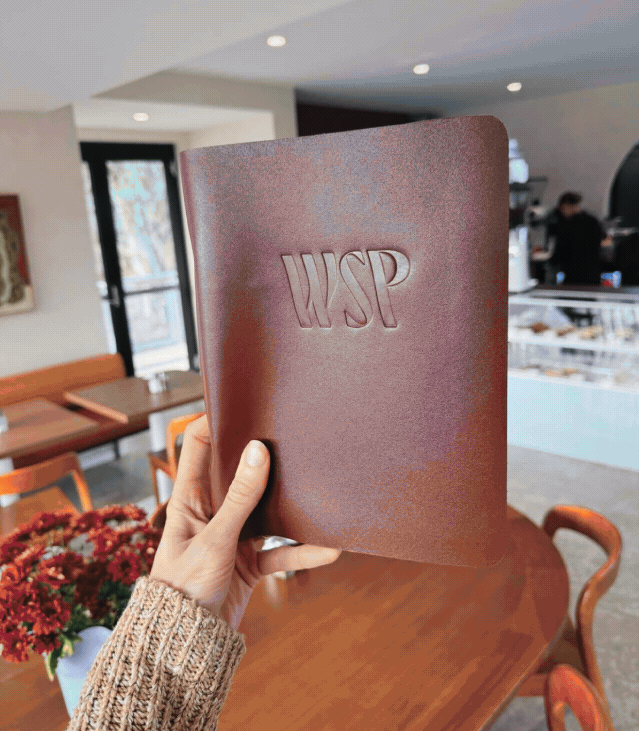 Person holding a maroon menu with embossed WSP lettering in a cozy cafe, with wooden tables, chairs, and a flower arrangement in the background.