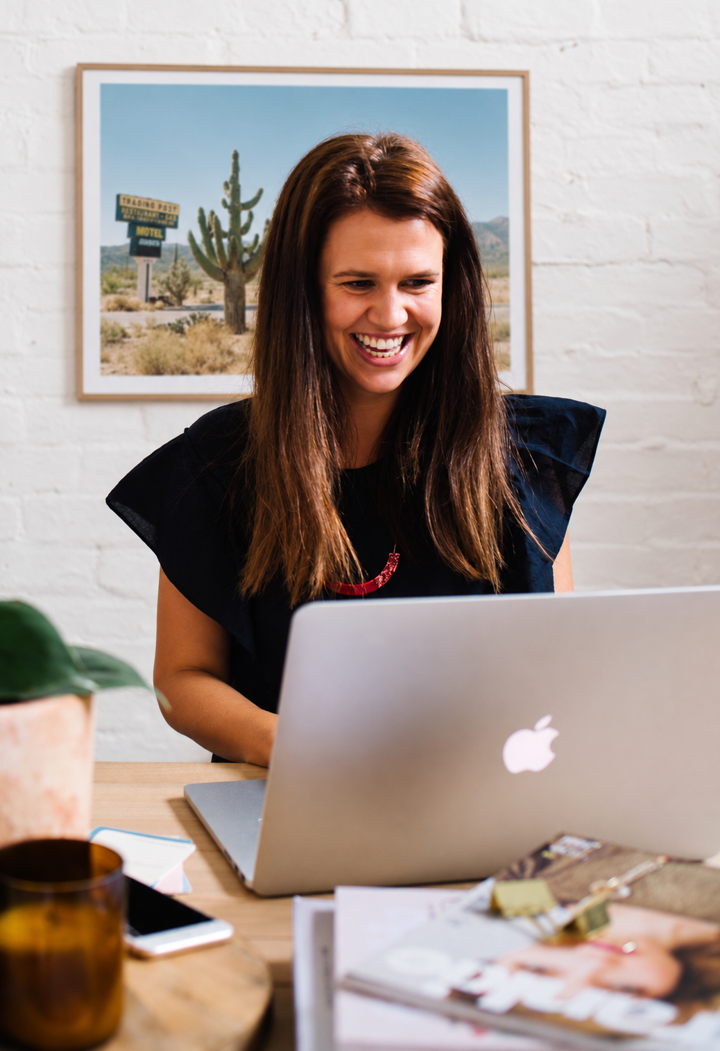 A woman with brown hair smiling while using a silver laptop at a wooden desk, with a brick wall and desert landscape photograph in the background.