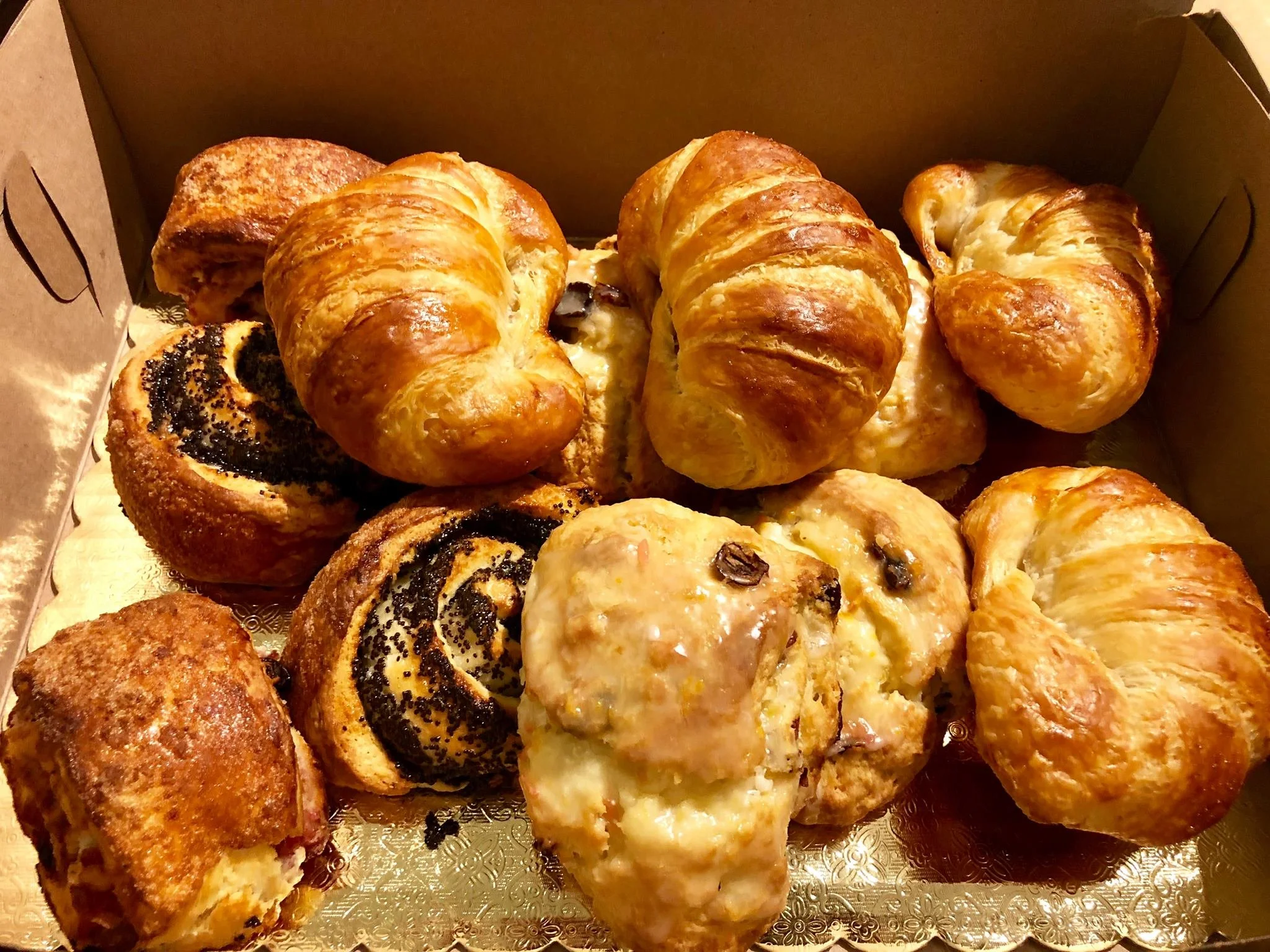 An assortment of baked pastries including croissants, cinnamon rolls, and glazed pastries in a cardboard box.