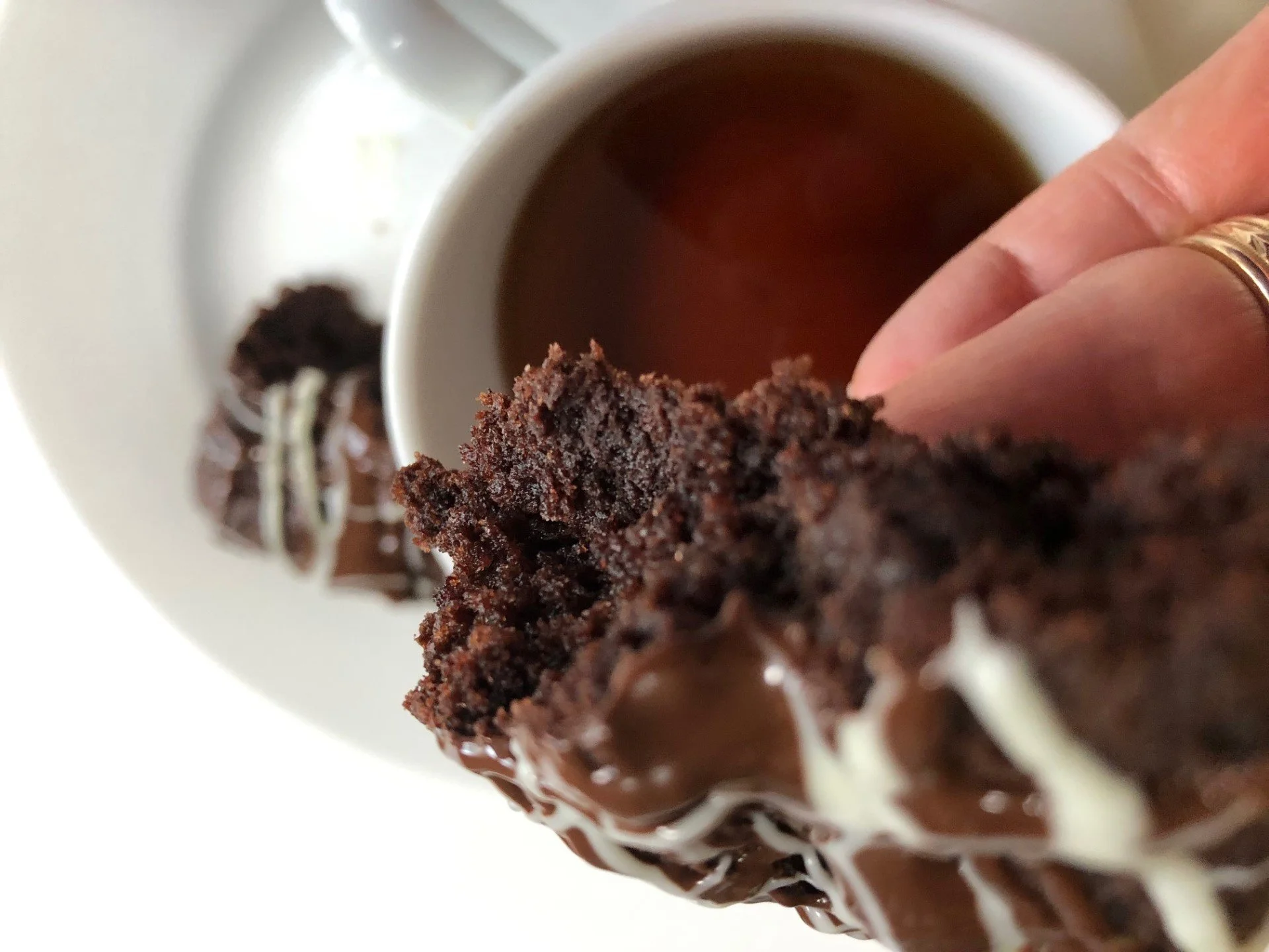 Close-up of a person holding a chocolate cookie with a cup of tea in the background on a white saucer.