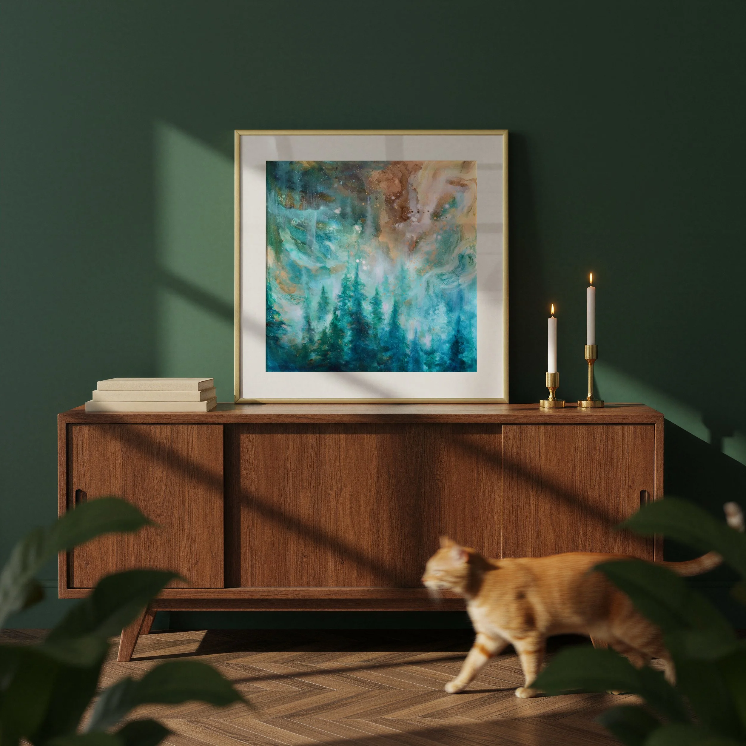 A wooden sideboard with a framed abstract painting of trees on a green wall, two tall candles in gold holders, some stacked books, and a blurred orange tabby cat in the foreground.