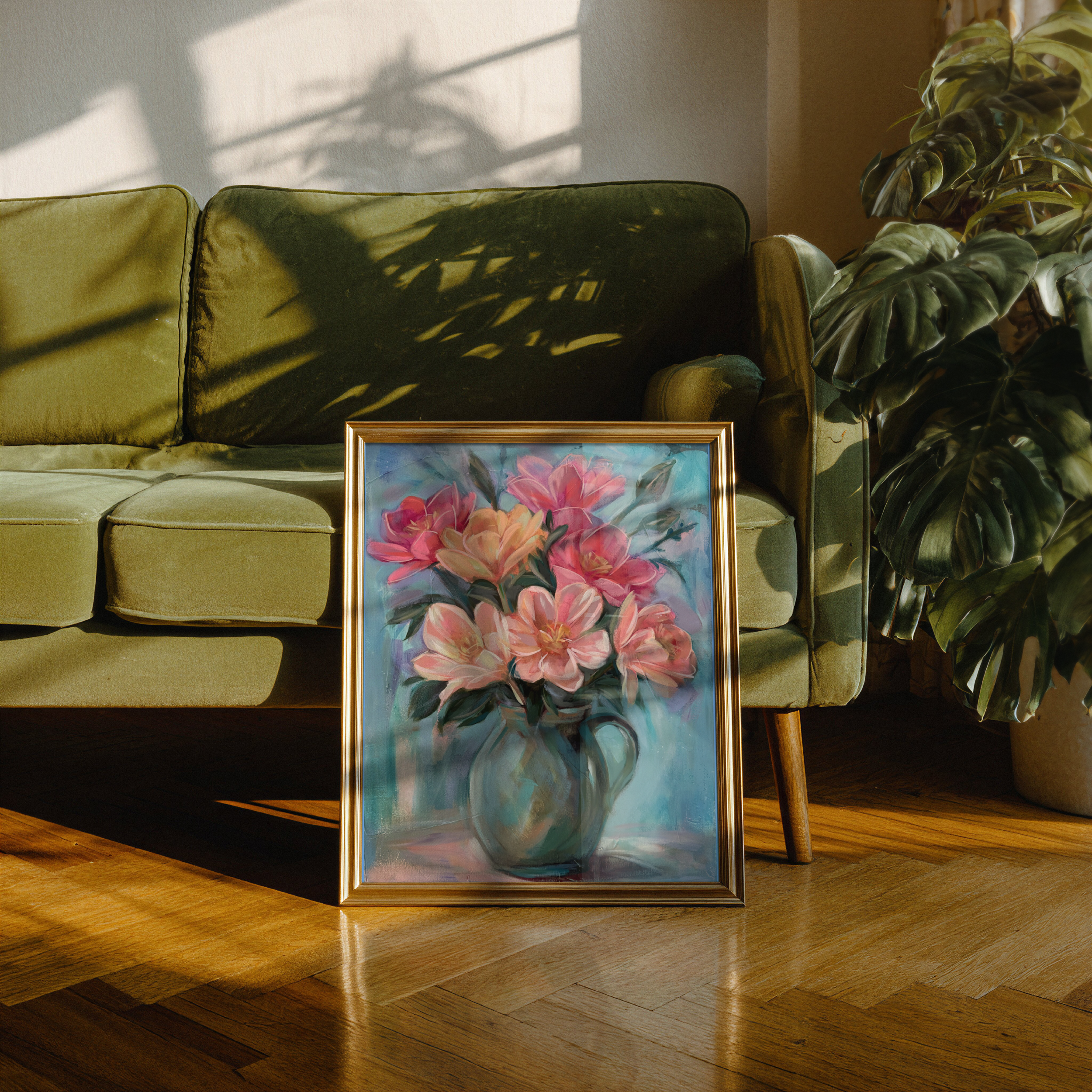 Painting of pink and orange flowers in a gold frame, leaning against a green velvet sofa with shadows of a window and a plant on the wall and sofa, on a wooden floor.