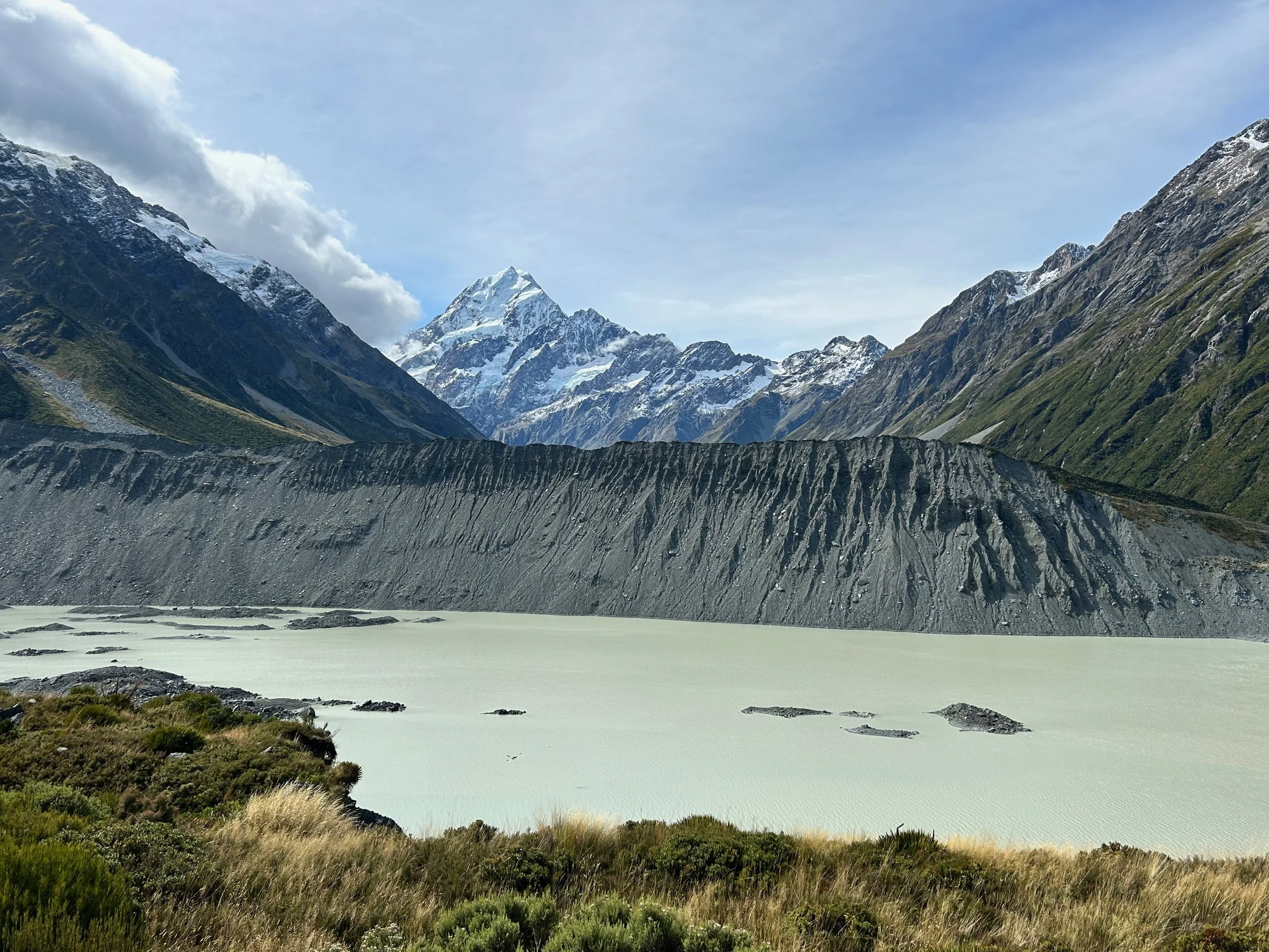 Scenic view of a mountain landscape with snow-capped peaks, rocky cliffs, a green valley, and a calm lake in the foreground.