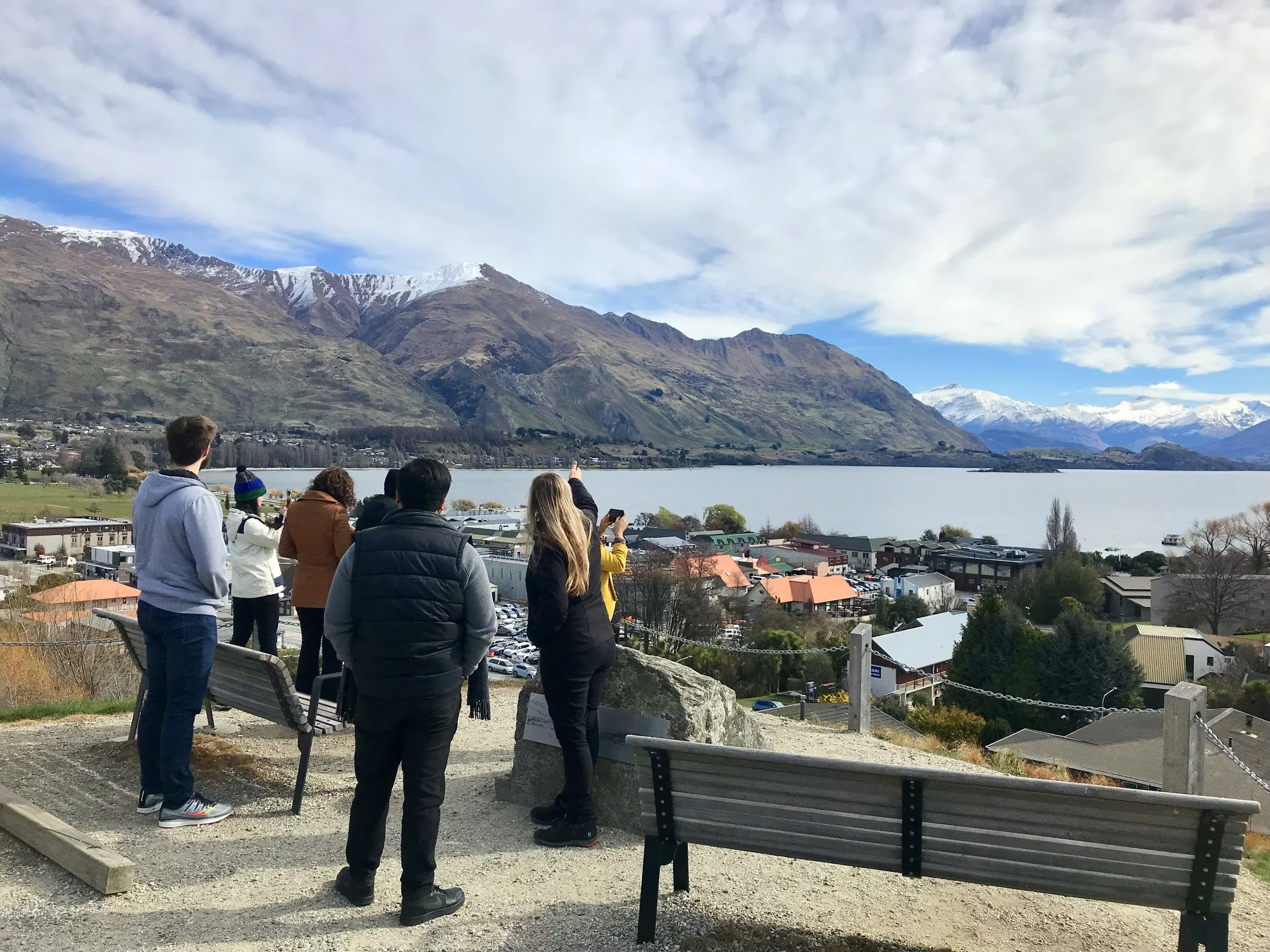 A group of people standing on an overlook, viewing a scenic landscape featuring a lake, mountains, and a small town with colorful rooftops. Snow-capped peaks are visible in the background under a partly cloudy sky.