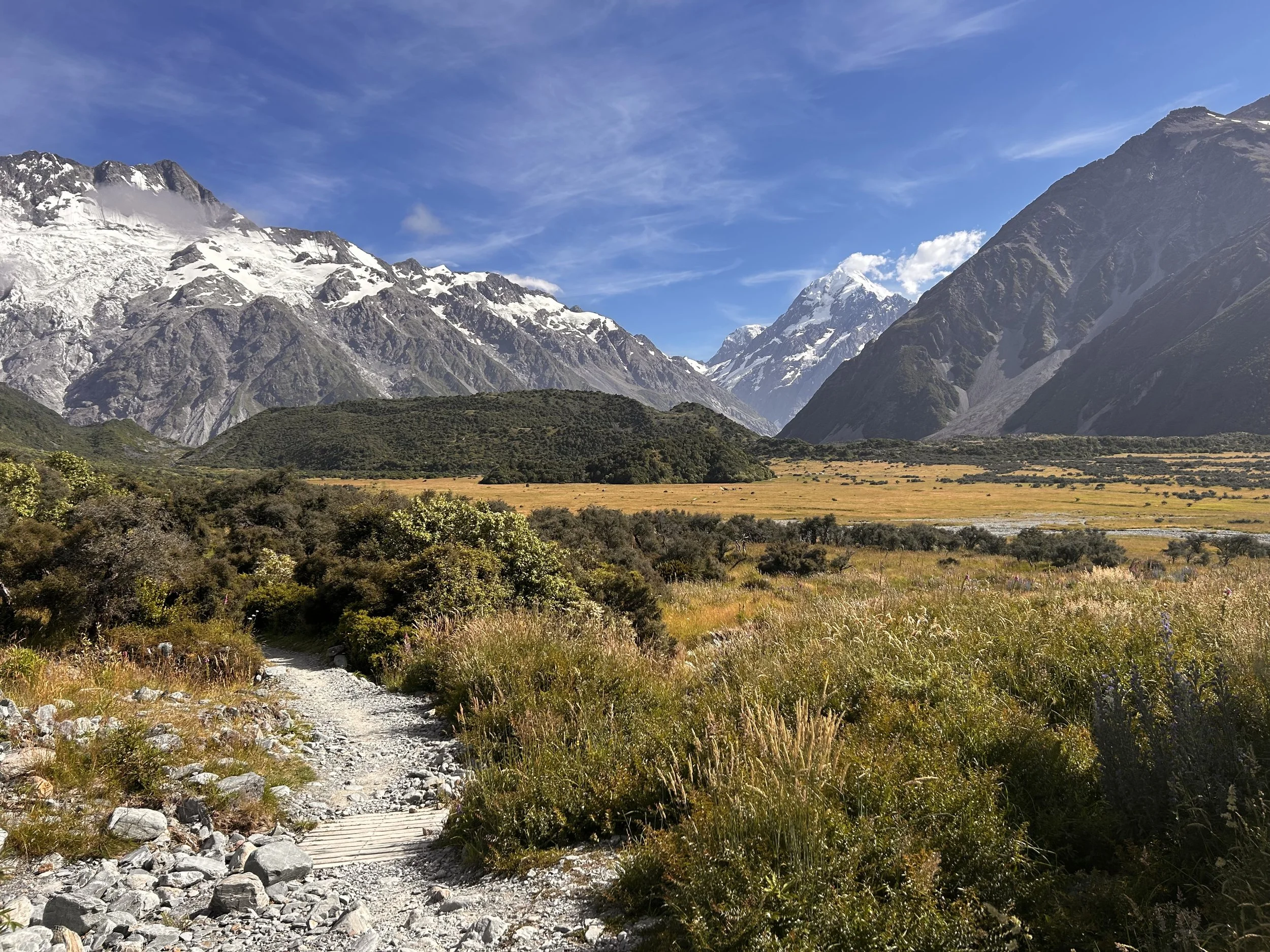 Mountain landscape with a rocky path, grassy fields, and snow-capped peaks under a partly cloudy sky.