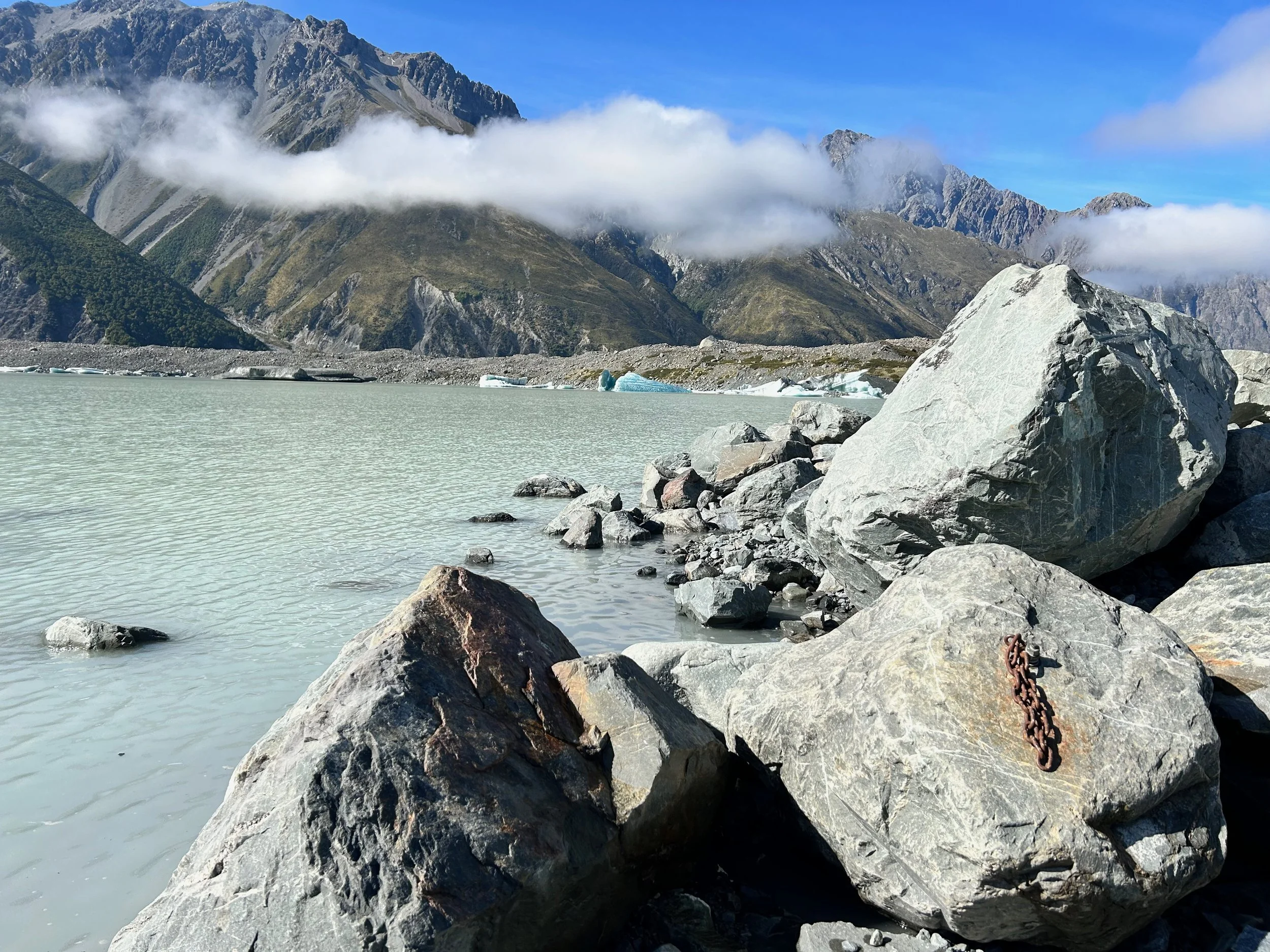Rocky lakeshore with large boulders, glacial lake, and distant mountain range under a blue sky with clouds.