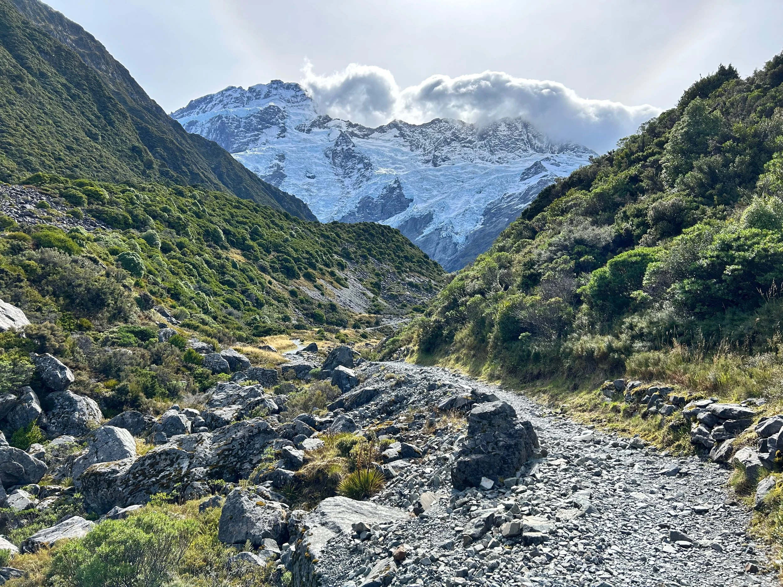 Mountain landscape with a rocky trail, lush green vegetation, and snow-covered peaks under a cloudy sky.