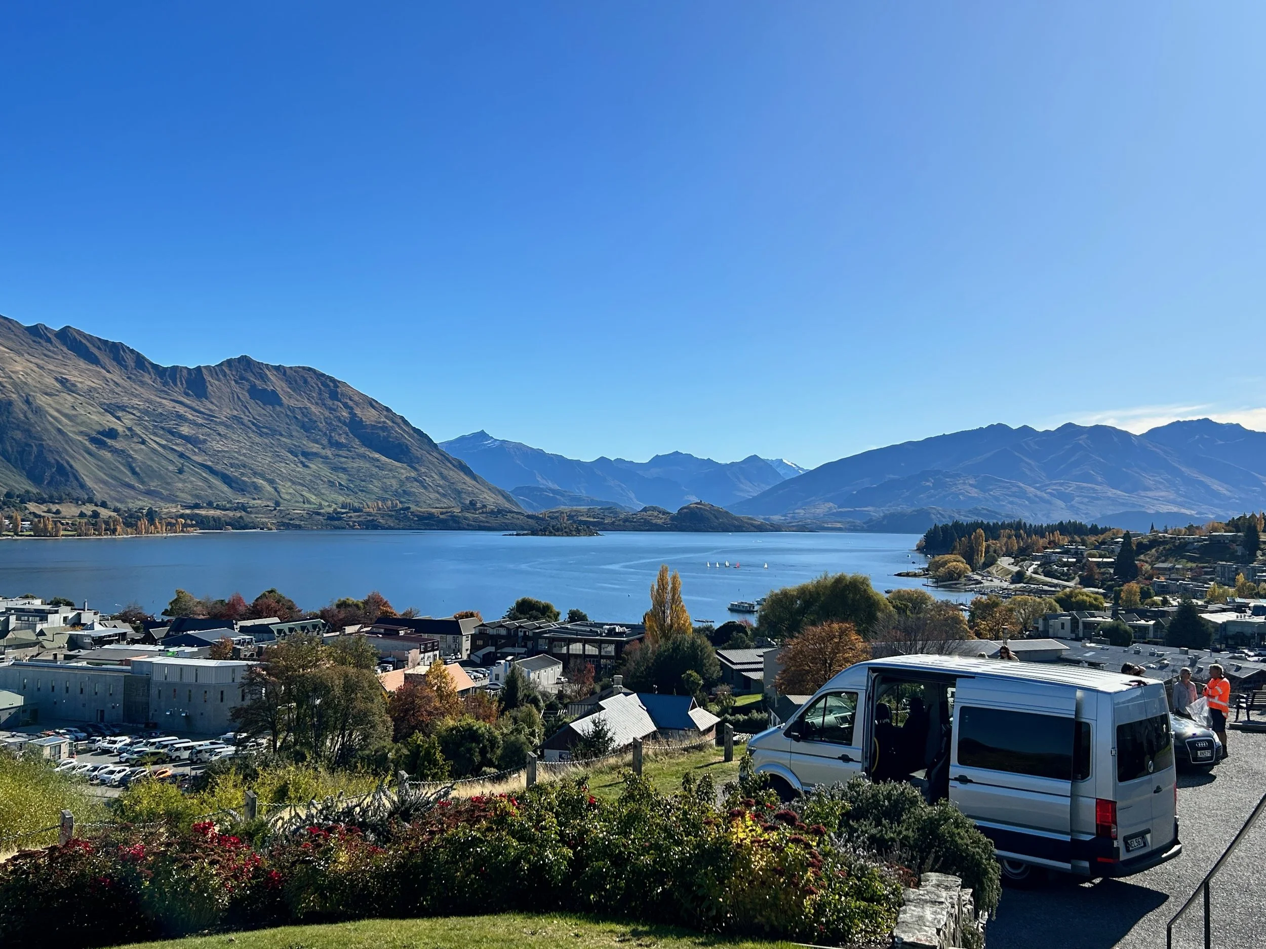 Panoramic view of a scenic Lake Wānaka with surrounding mountains, a town, and a van parked in the foreground.