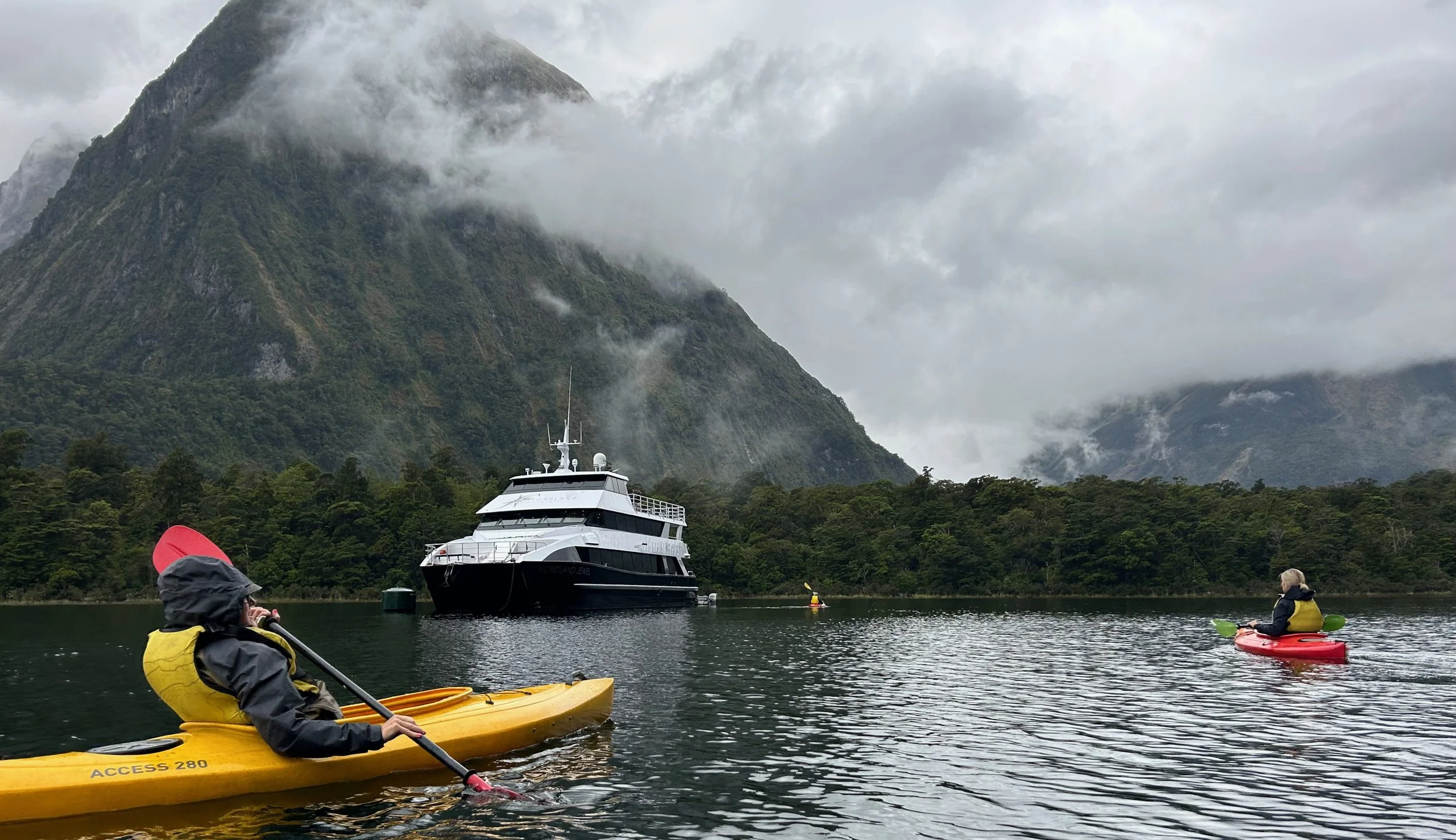 Two people kayaking on a lake with a large tour boat nearby and a misty mountain in the background.