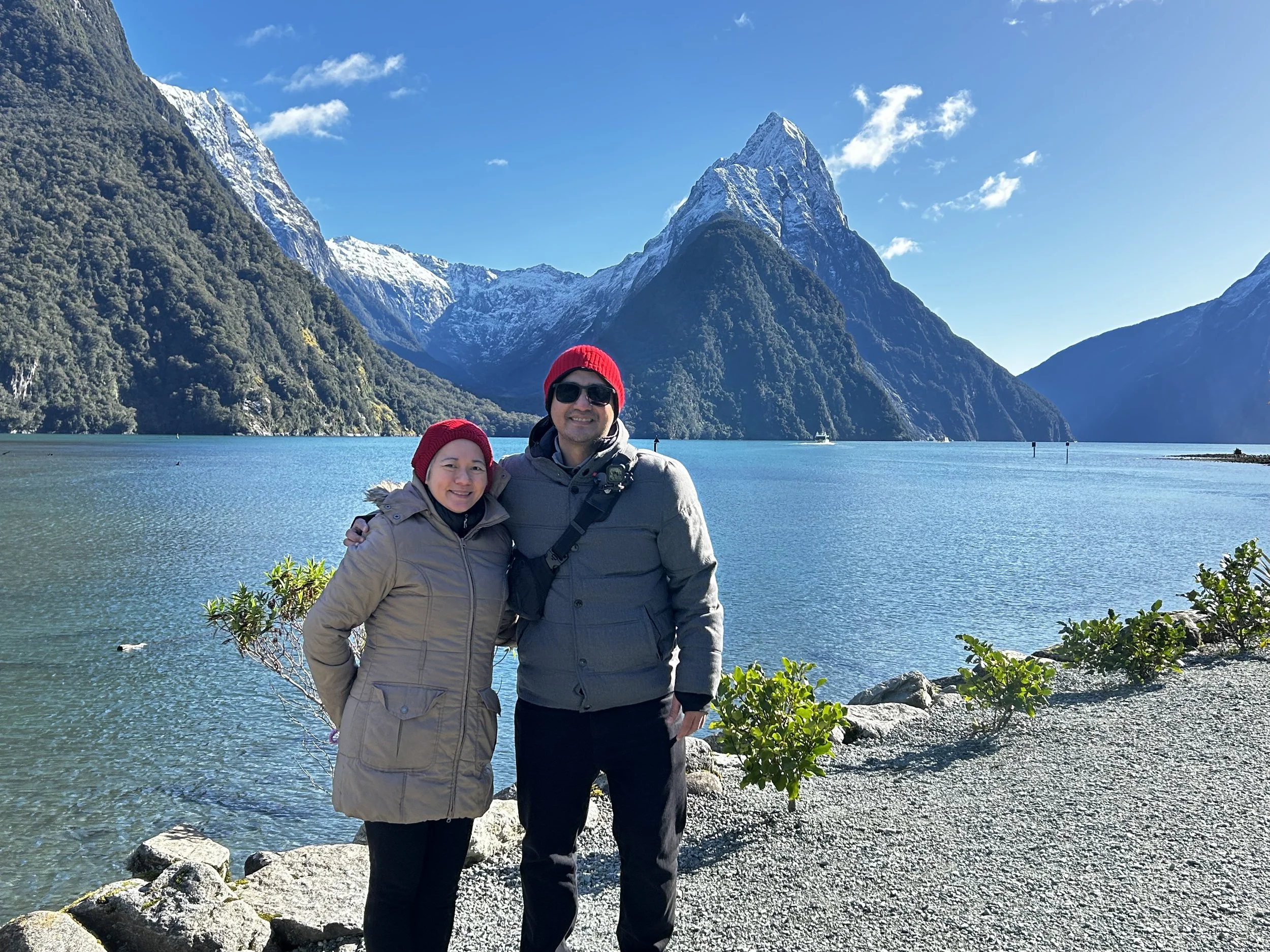 Two people wearing winter clothing and red hats standing in front of Mitre Peak Fiordland