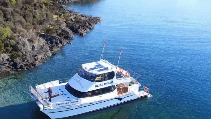 A tour boat named "Dual Image" is on a tranquil lake, near a rocky forested shoreline. It has flags and life rings, with "Daily Lake Cruises" written on its side.