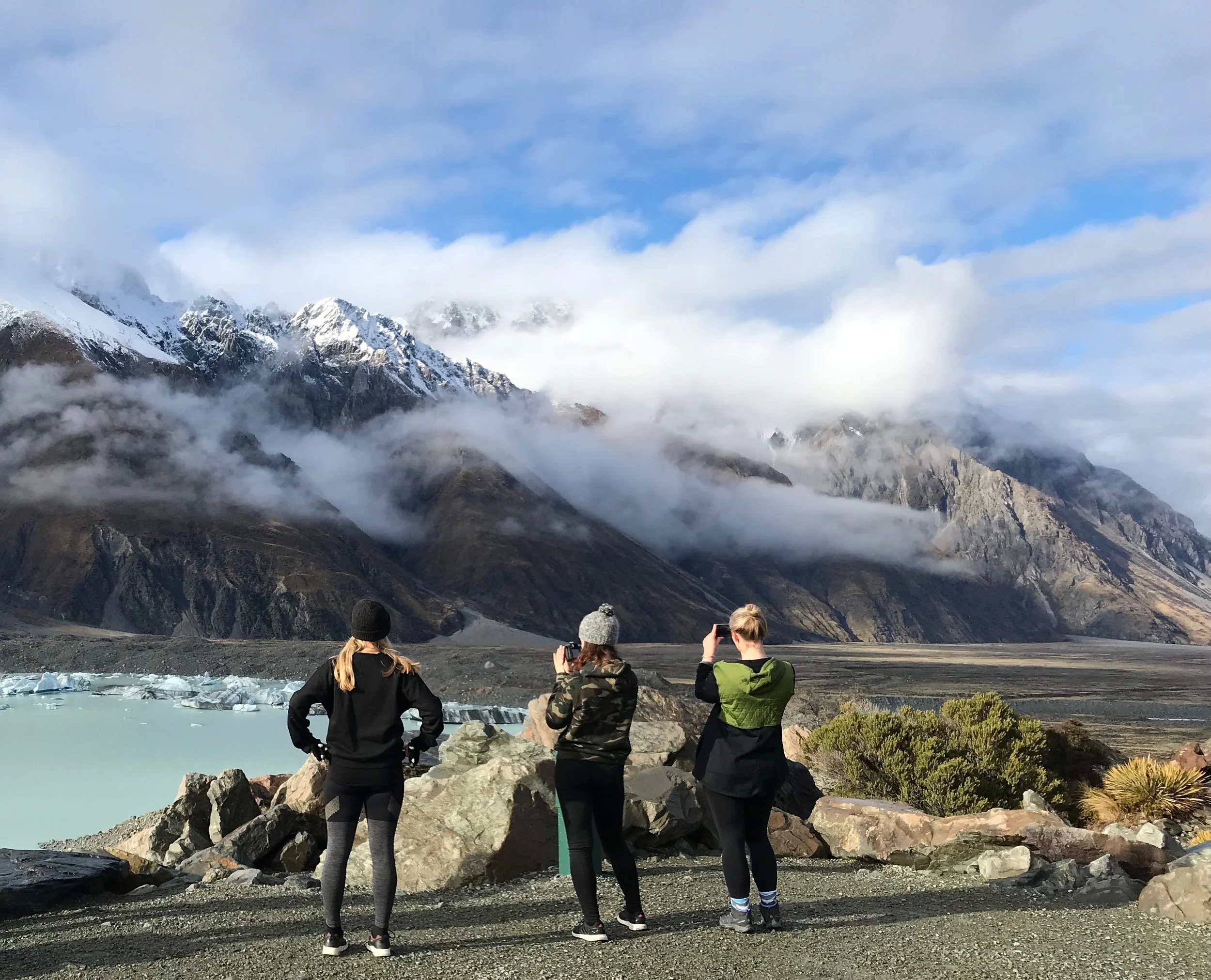 Three people stand on rocky ground, facing snowy mountains and a lake under cloudy skies.