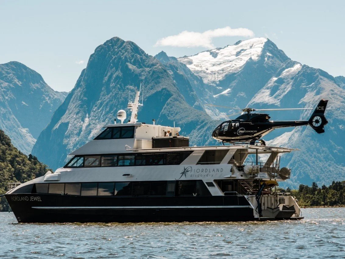 Luxury yacht named "Fiordland Jewel" with a helicopter on deck, set against a backdrop of snow-capped mountains and water.