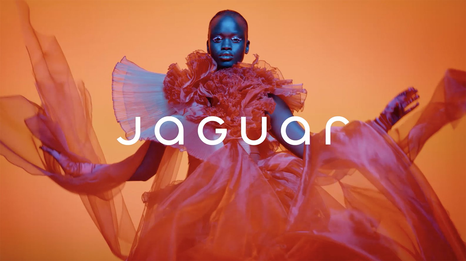 Jaguar logo over a photograph of a women in a red eclectic dress on orange studio background
