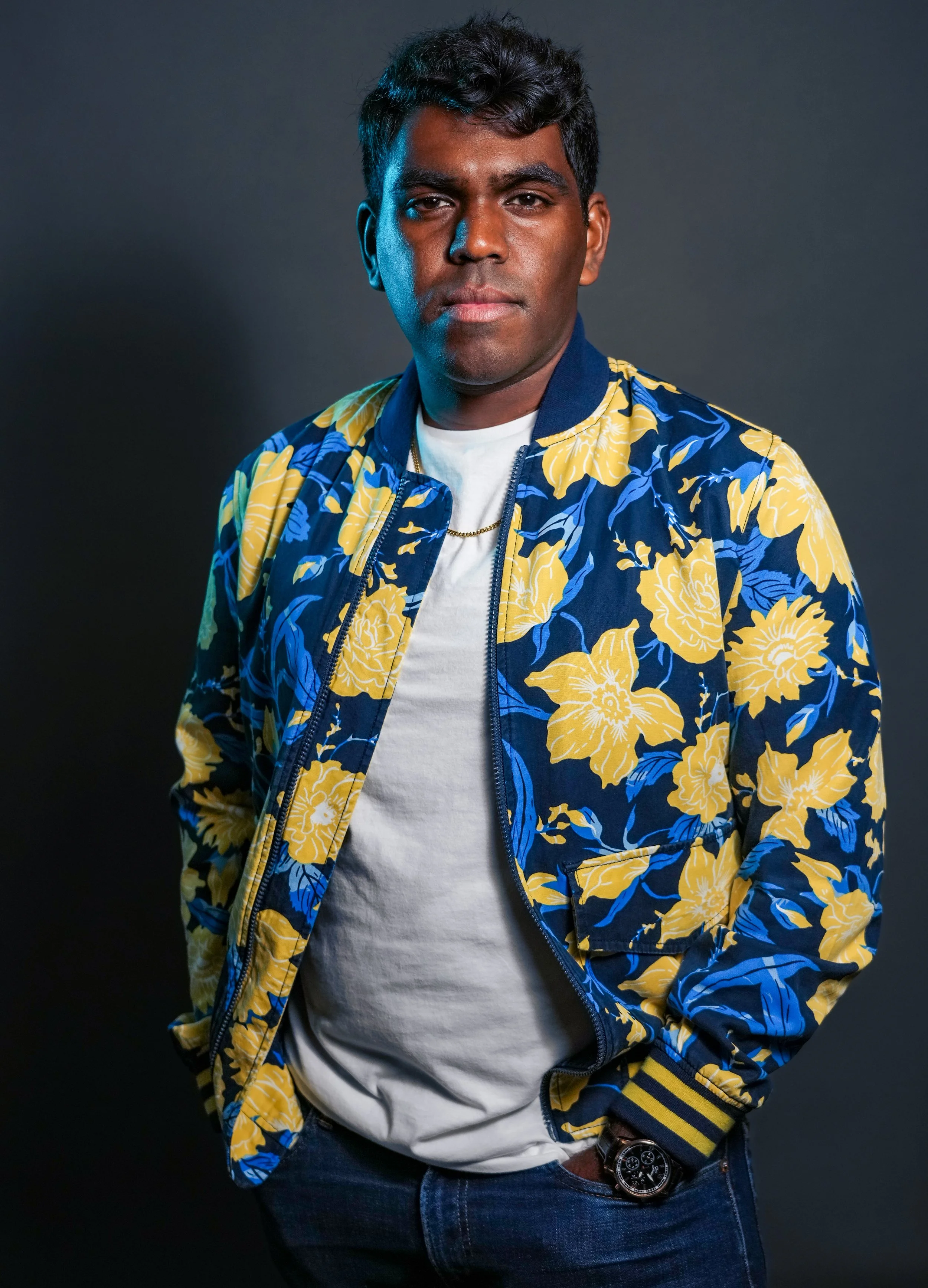 A young man with dark hair and medium skin wearing a colorful blue and yellow floral bomber jacket over a white t-shirt, posing against a dark background.