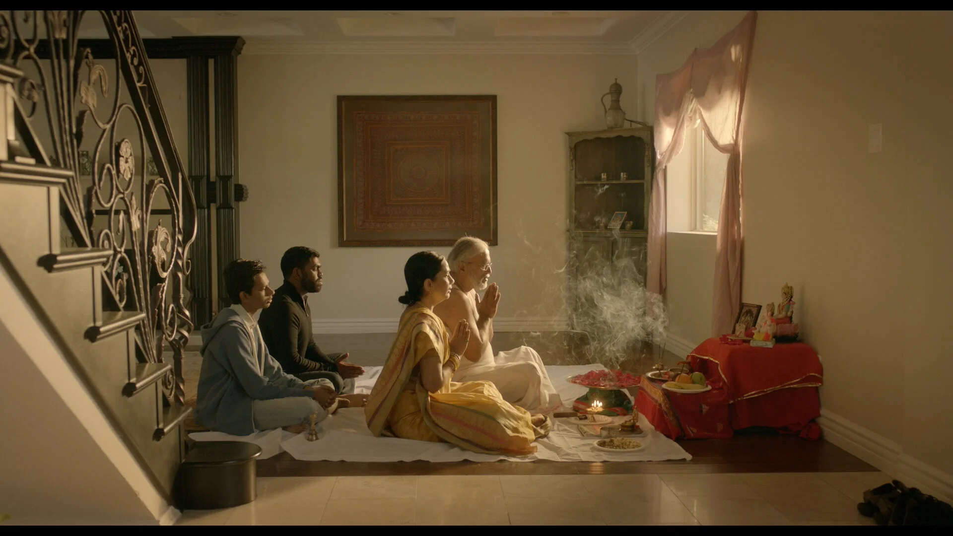 Four people sitting on the floor in a prayer pose during a Hindu religious ceremony in a living room, with incense smoke rising.