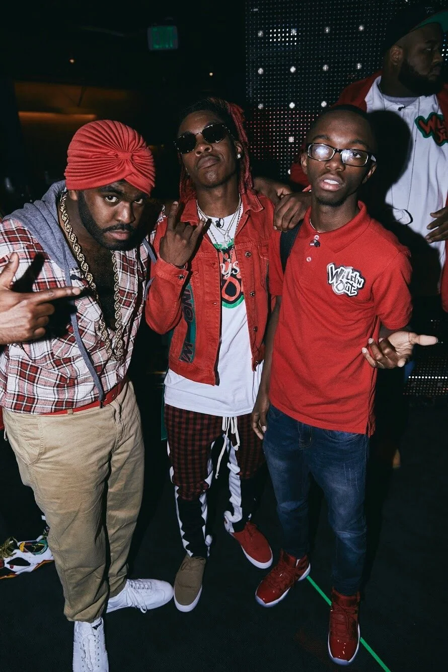 Three men posing together in a nightclub with a dark background and LED lights. Two of them are making hand gestures, one with sunglasses and red-tied hair, and the other with glasses and a red polo shirt.
