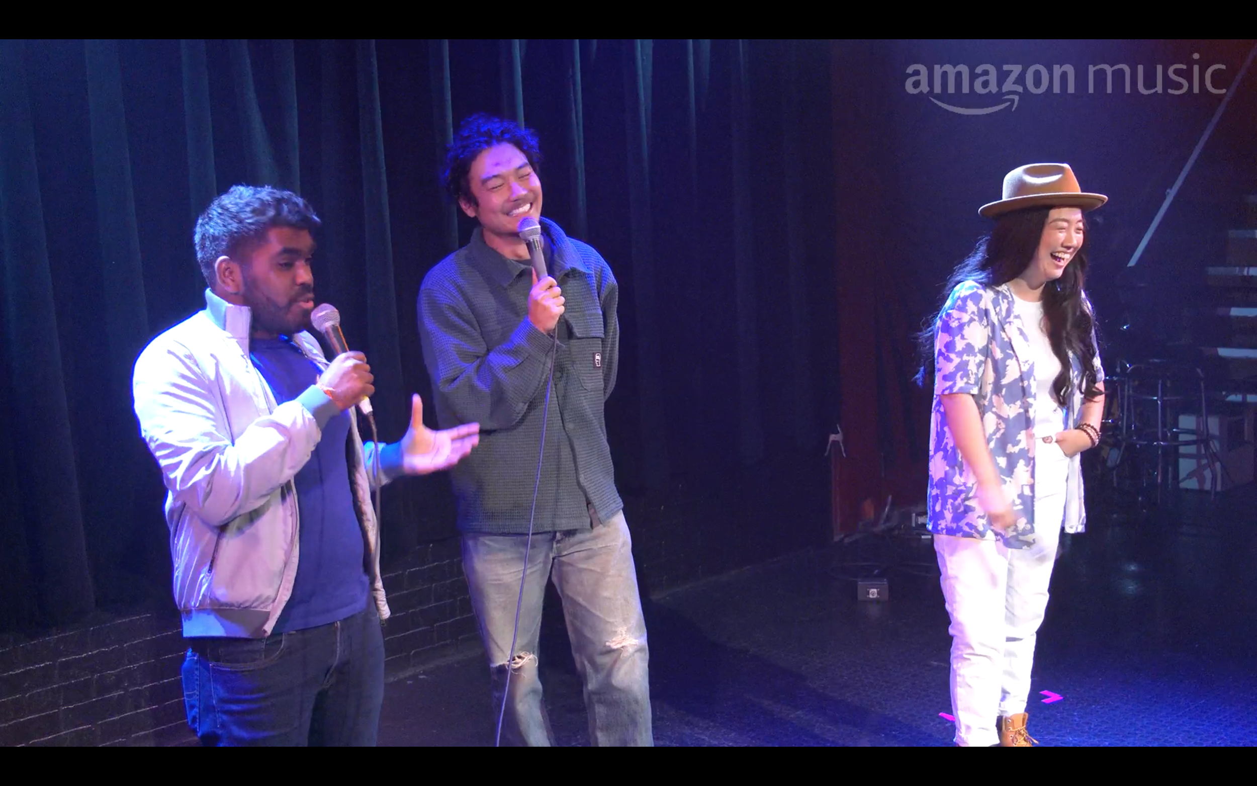 Three people standing on a stage, each holding a microphone, laughing and engaging with the audience against a dark curtain backdrop, with the Amazon Music logo visible in the top right corner.