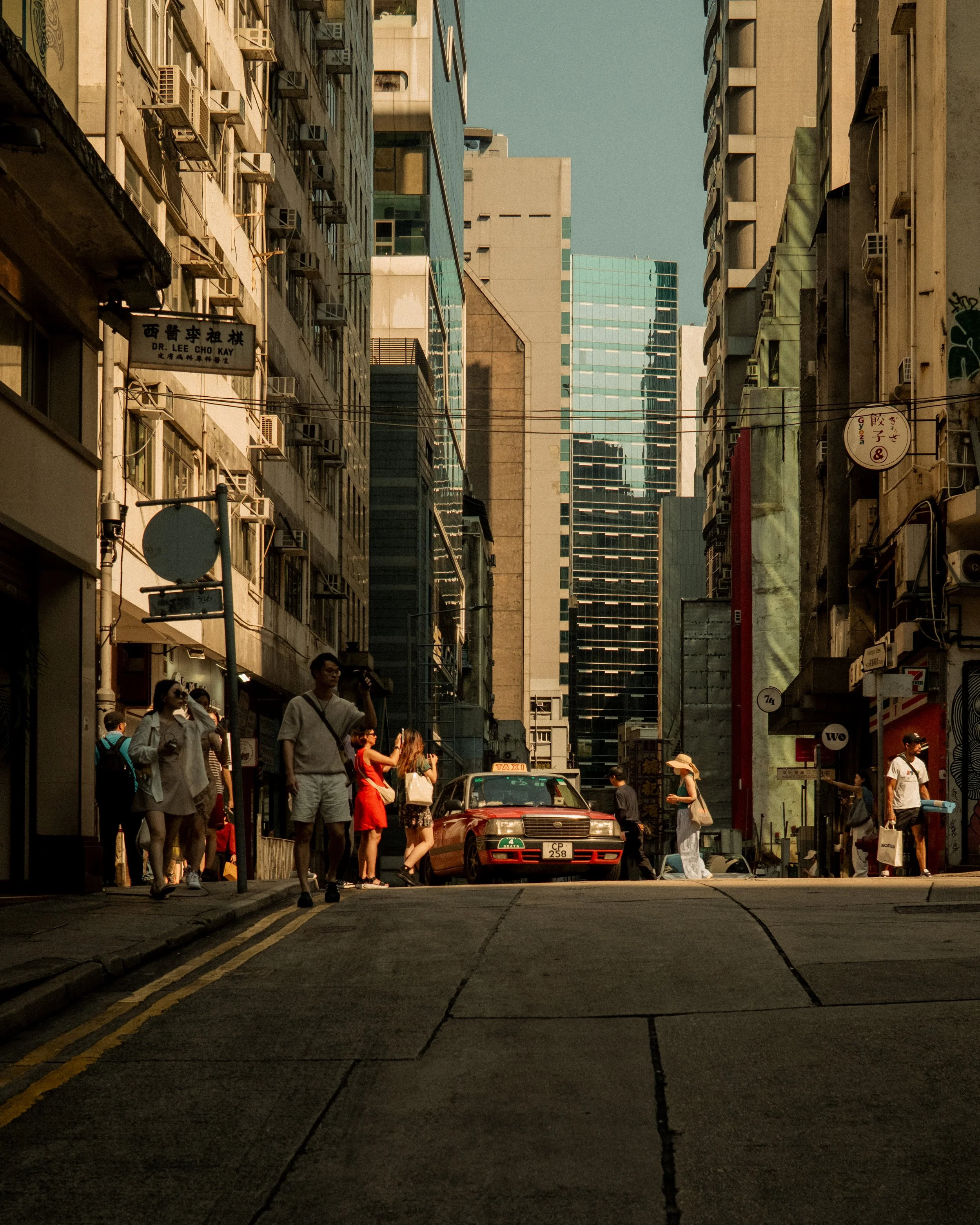 City street scene with pedestrians crossing in front of a red taxi, surrounded by tall buildings with varied architectural styles.