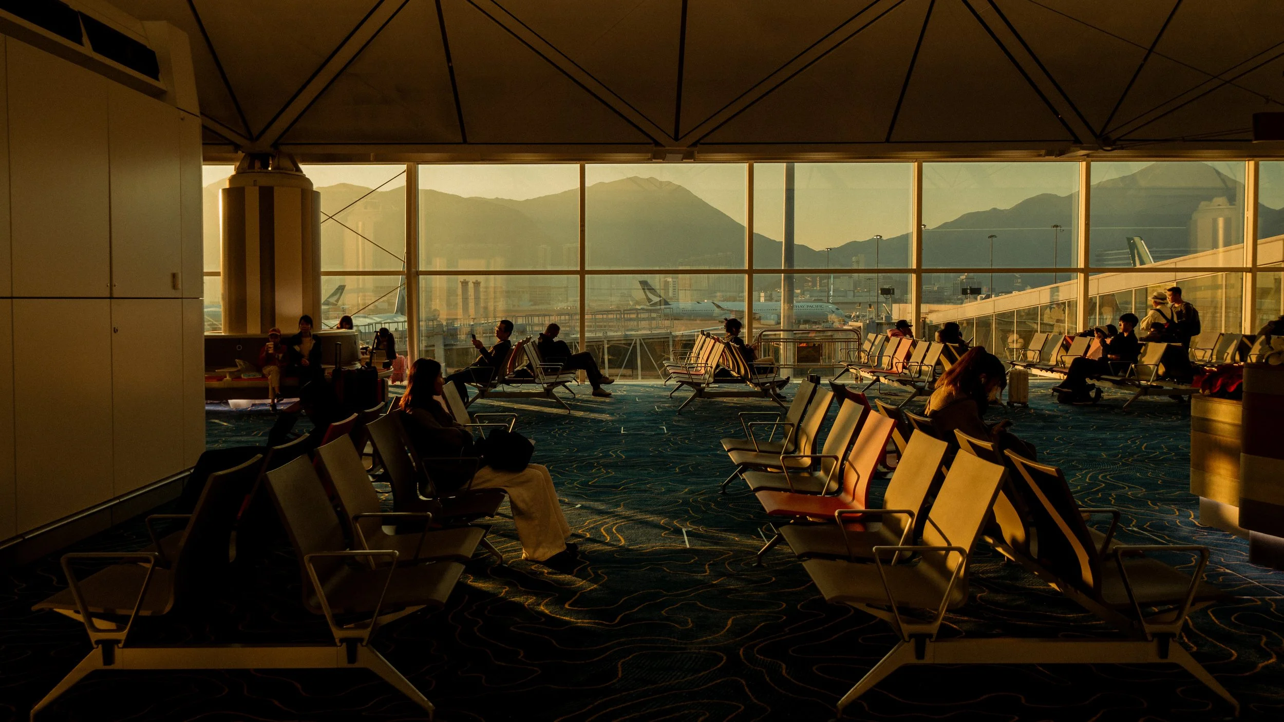 View of an airport terminal interior with seated travelers and large windows showing planes and mountains outside during sunset.