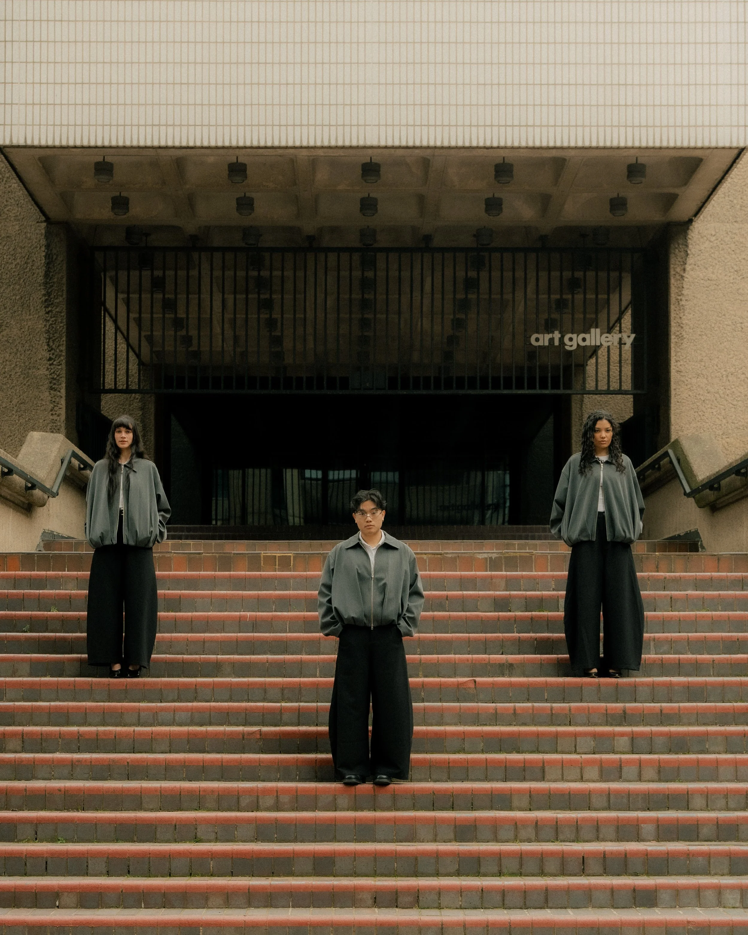 Three young individuals standing on a staircase outside of an art gallery building with a modern architectural design. They are wearing matching gray jackets and black pants, looking serious. The photo is taken during the day.