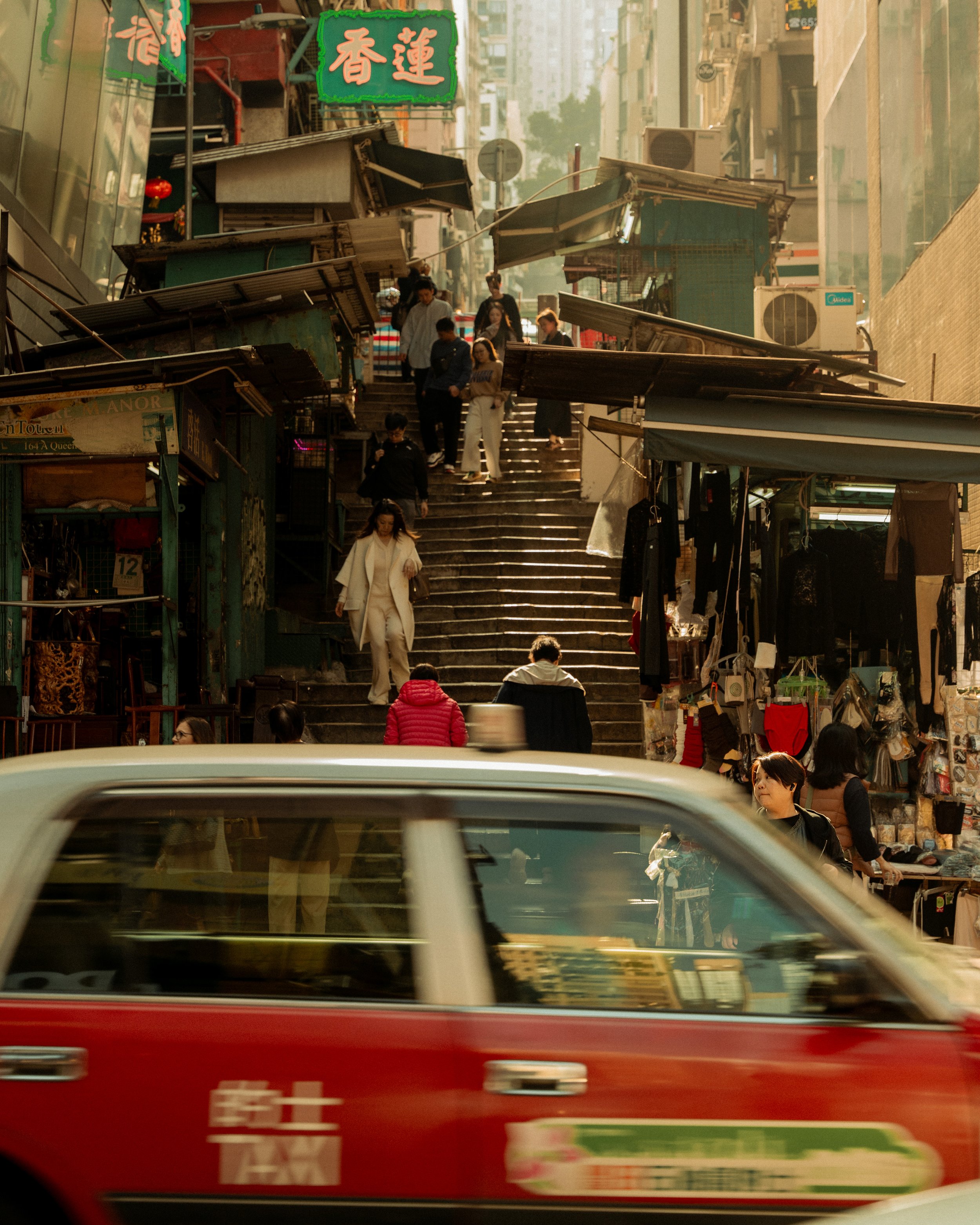 A busy street scene in an Asian city with a red taxi in the foreground and pedestrians walking on stairs and a marketplace lined with shops on either side.