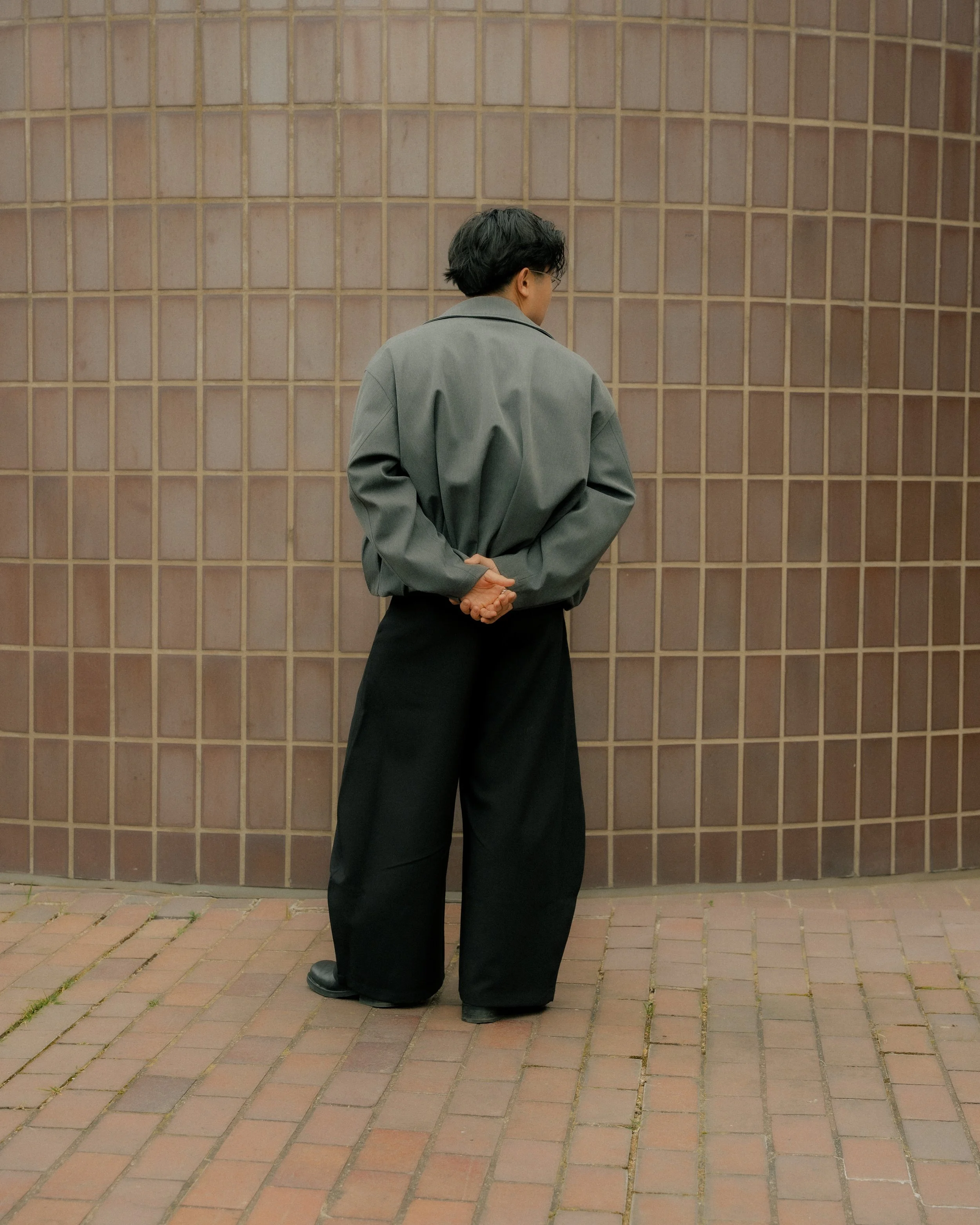 Person standing with their back turned, dressed in a gray jacket and black wide-leg pants, against a tiled wall.