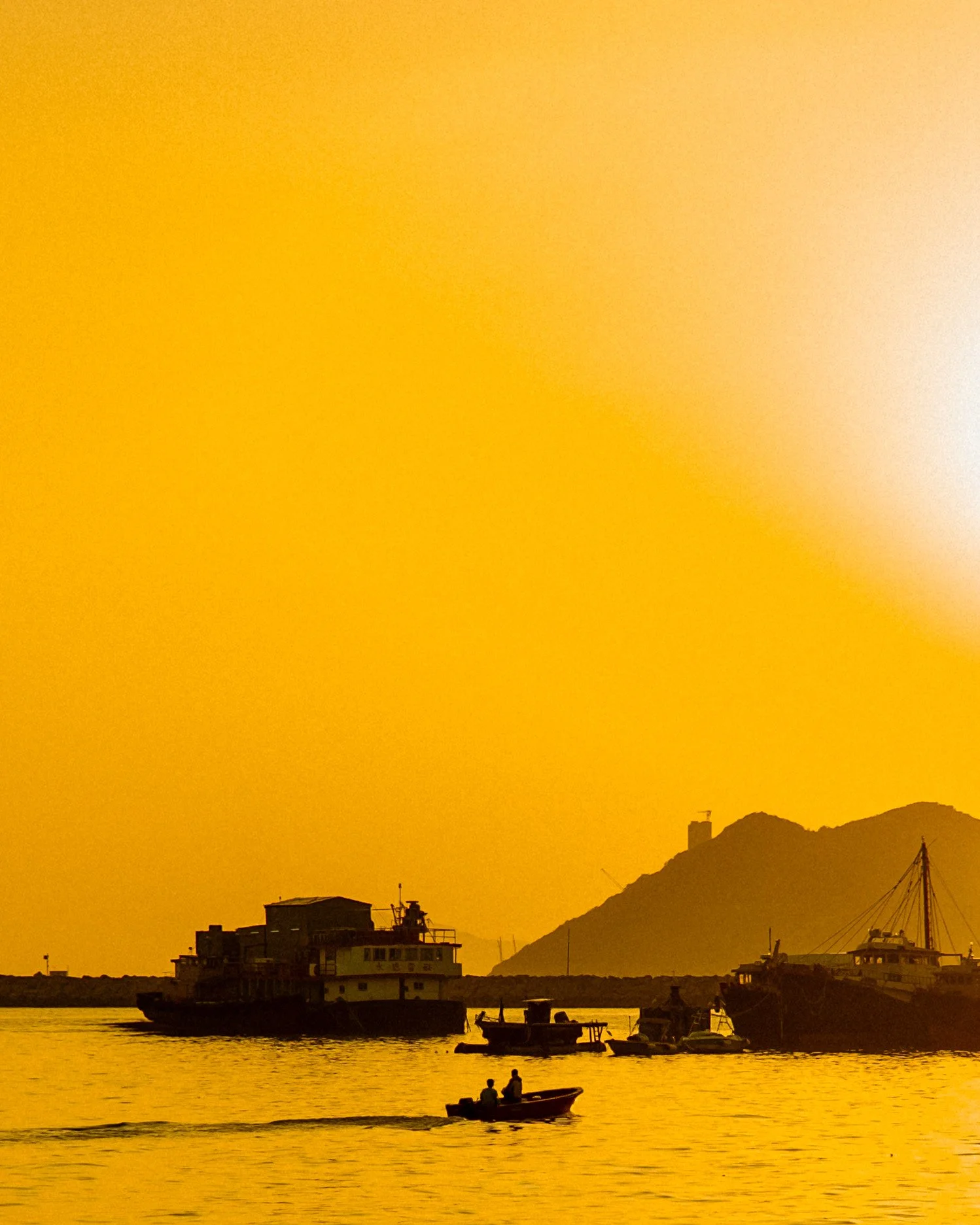 Sunset over a harbor with boats, including a large ship and smaller vessels, with a mountain in the background and the sky in golden hues.
