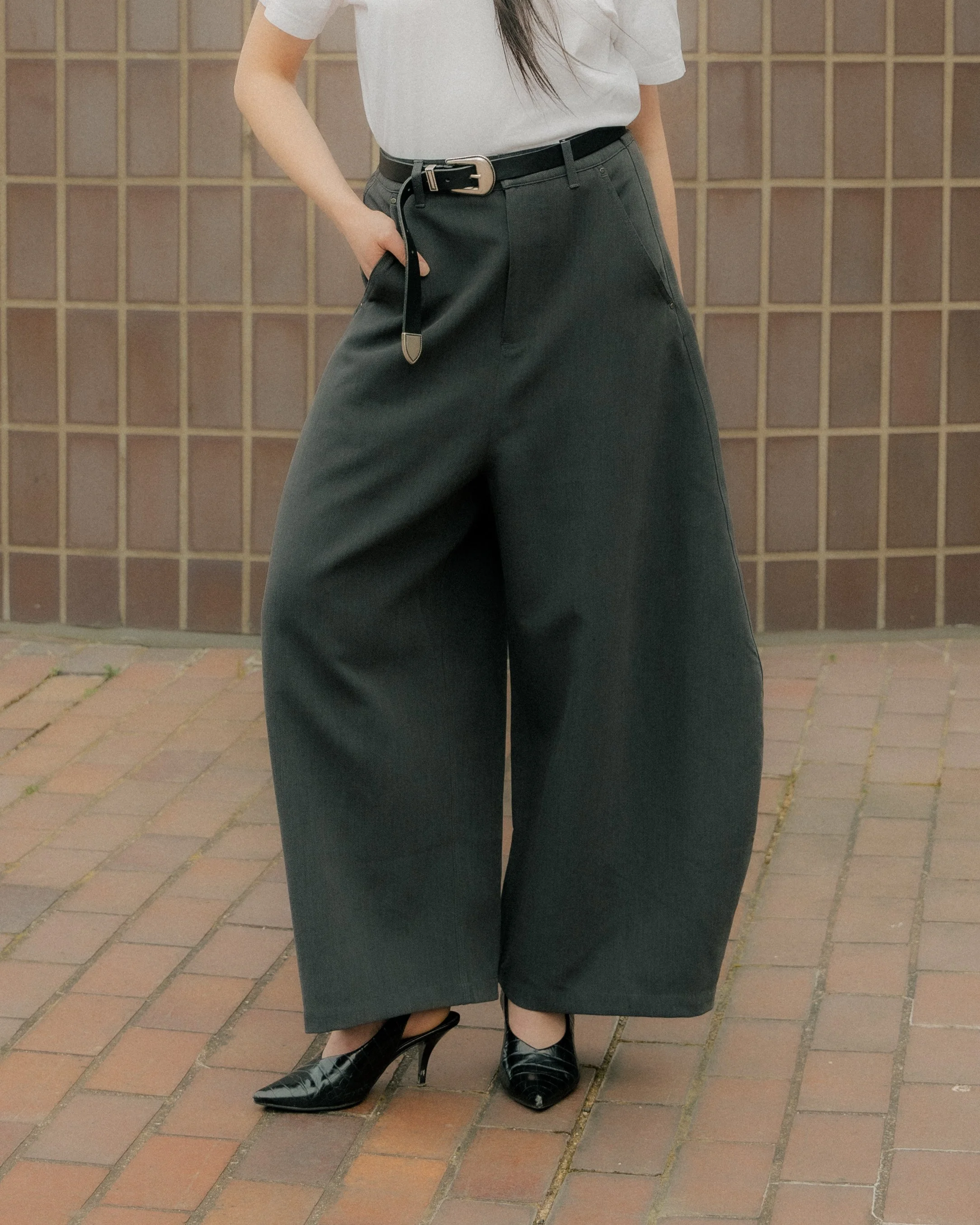 Person wearing a white shirt, black trousers with a belt, and black stiletto heels, standing on a brick sidewalk in front of a brown tiled wall.