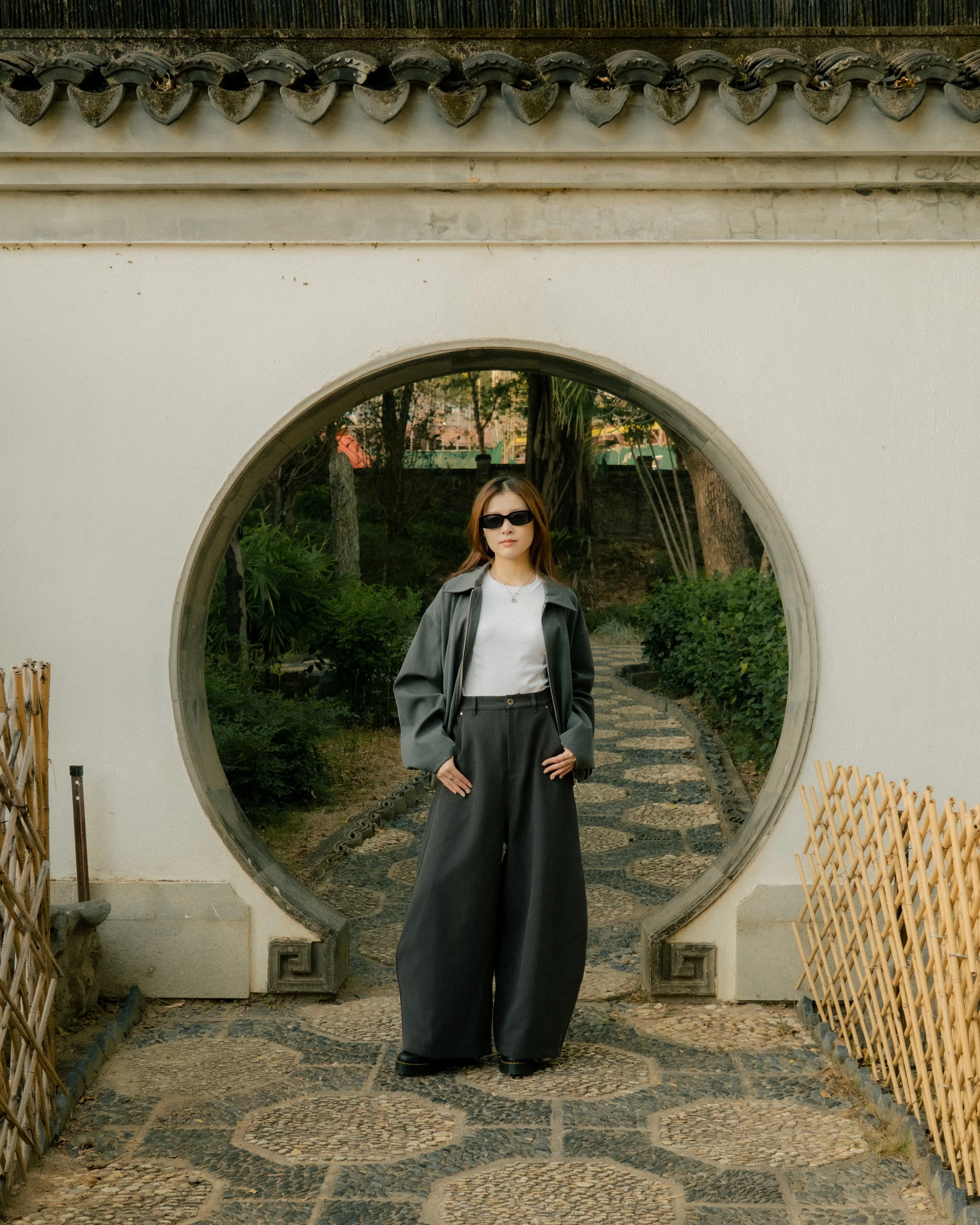Woman standing in front of a circular mirror arch, wearing sunglasses, a white shirt, and a gray jacket, with a garden background.