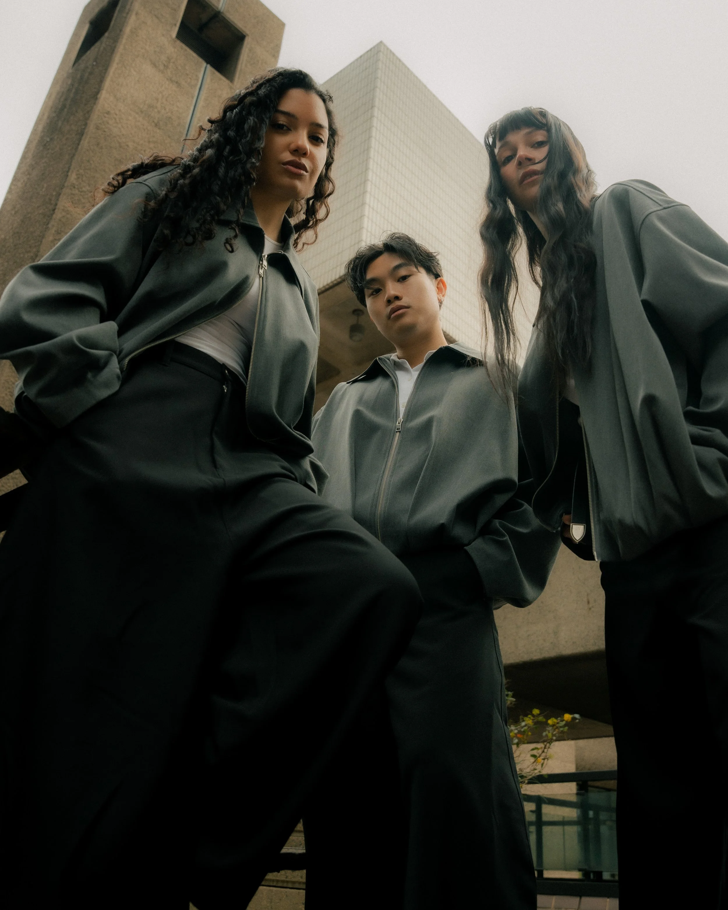 Three young adults looking down at the camera, standing outside between buildings with an overcast sky, wearing dark jackets.